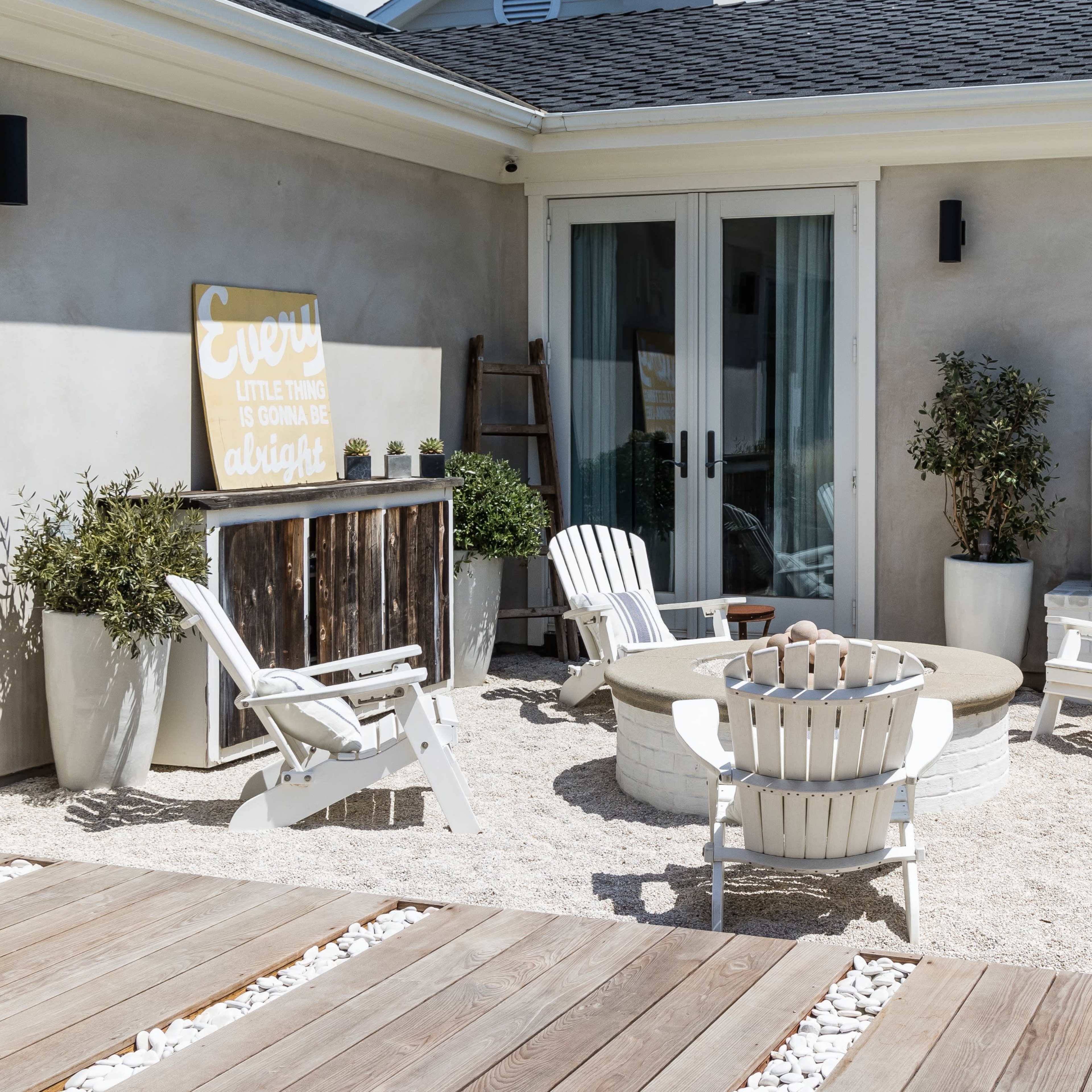 A small outdoor seating area with white chairs arranged around a circular fire pit, complemented by planters and a decorative sign on the wall.