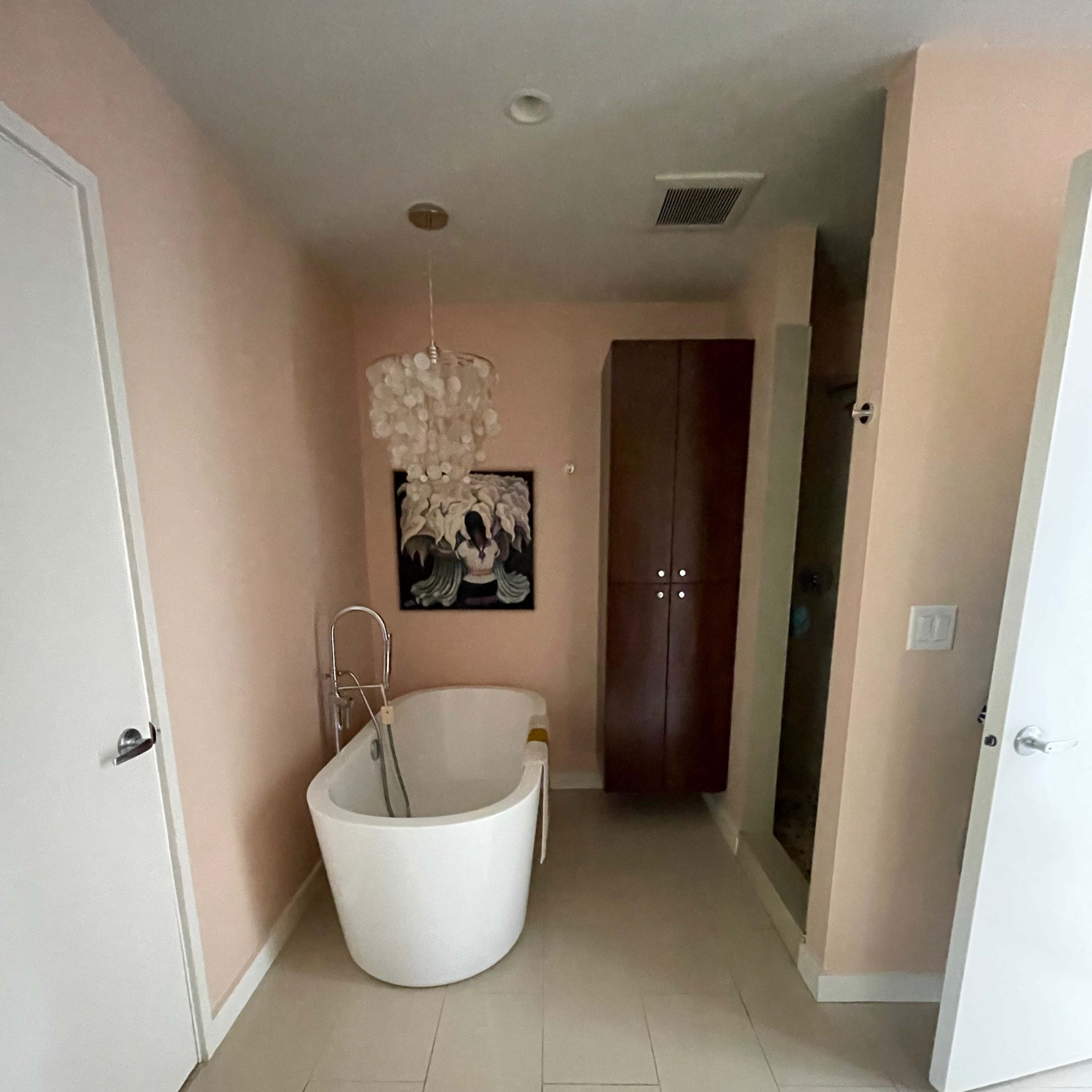 A bathroom with a freestanding white bathtub, a chandelier, light pink walls, a wooden cabinet, and a shower area.