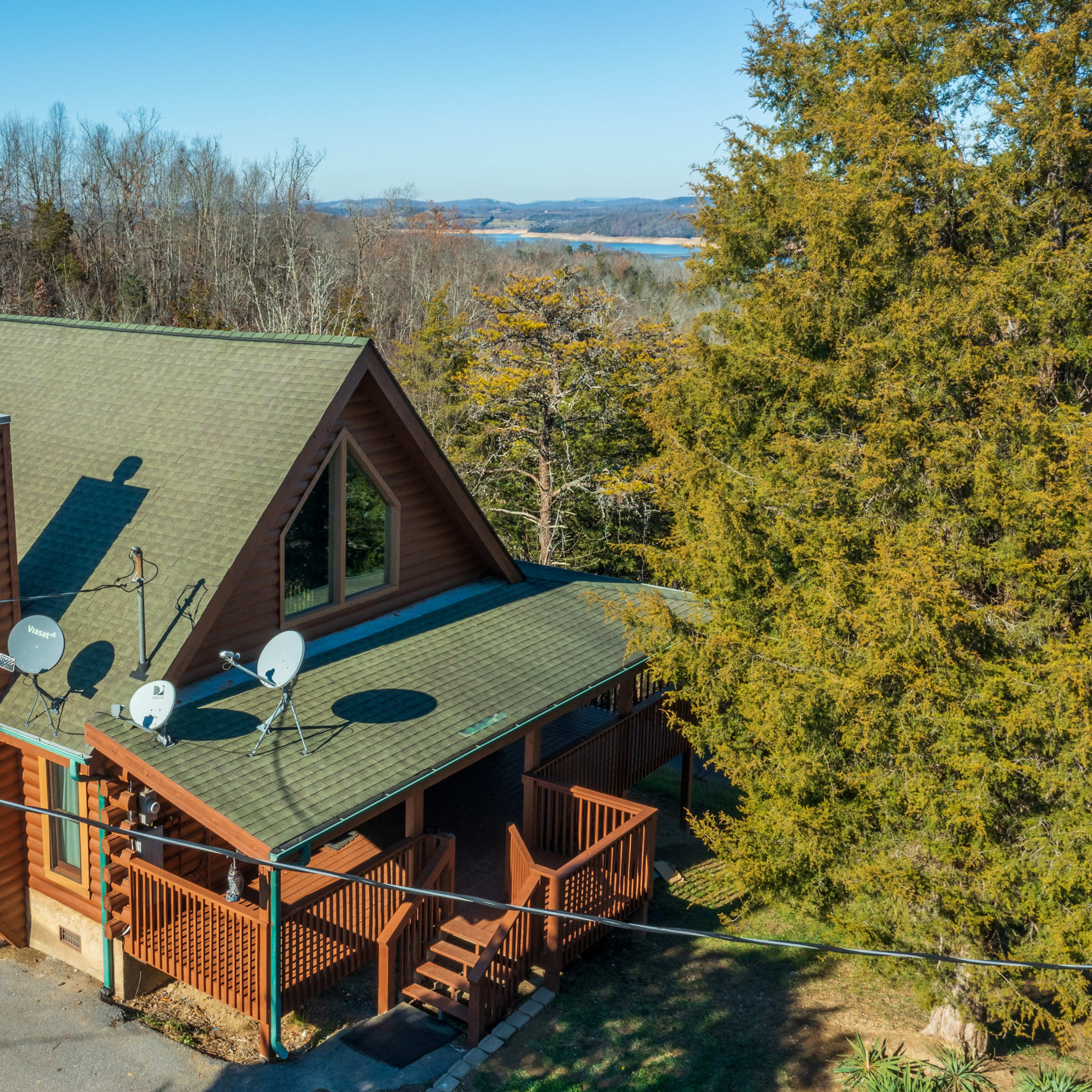 A wooden house with a sloped roof and satellite dishes sits surrounded by trees and overlooks a body of water in the distance.