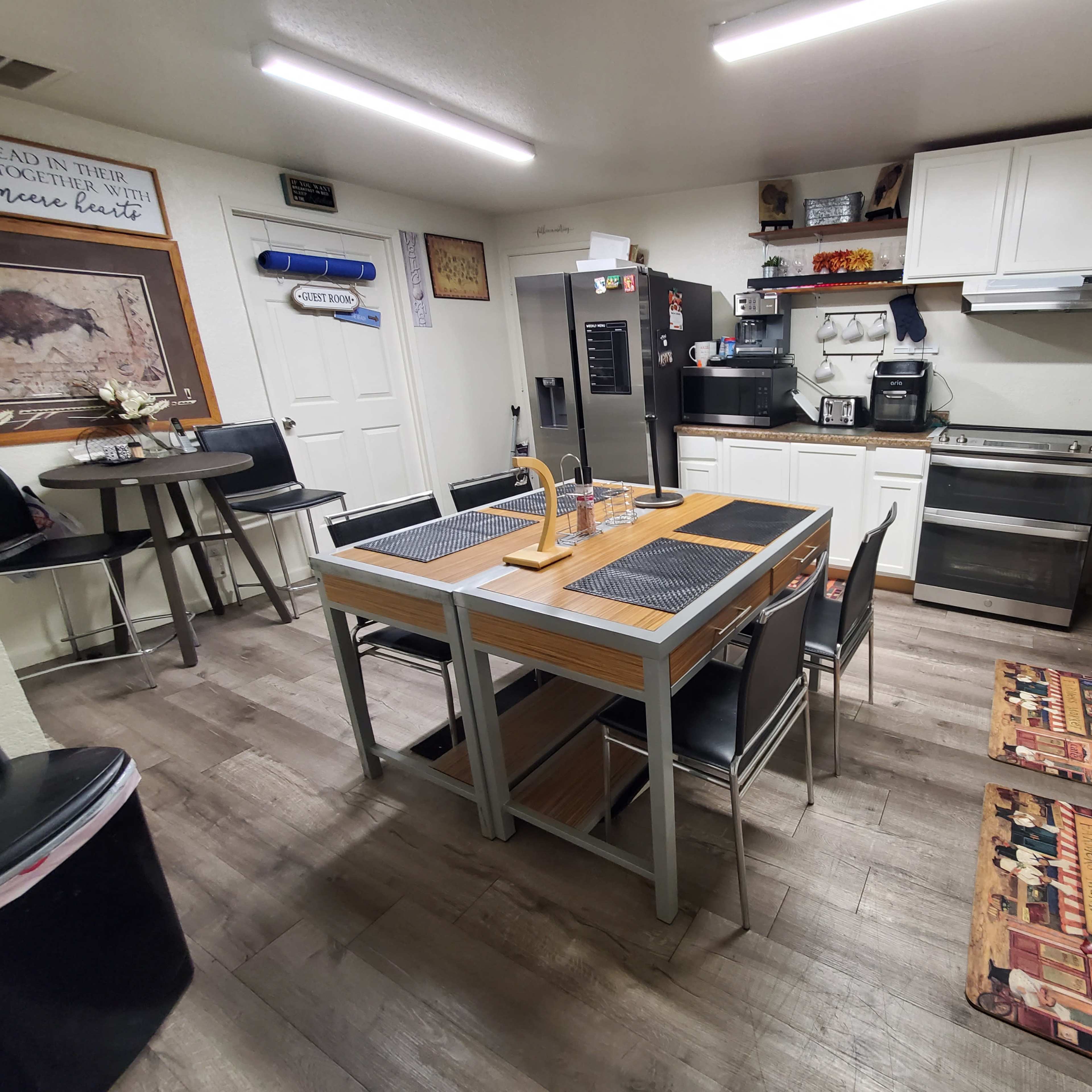 A modern kitchen with a central dining table, four metal chairs, a stainless steel refrigerator, and white cabinetry.