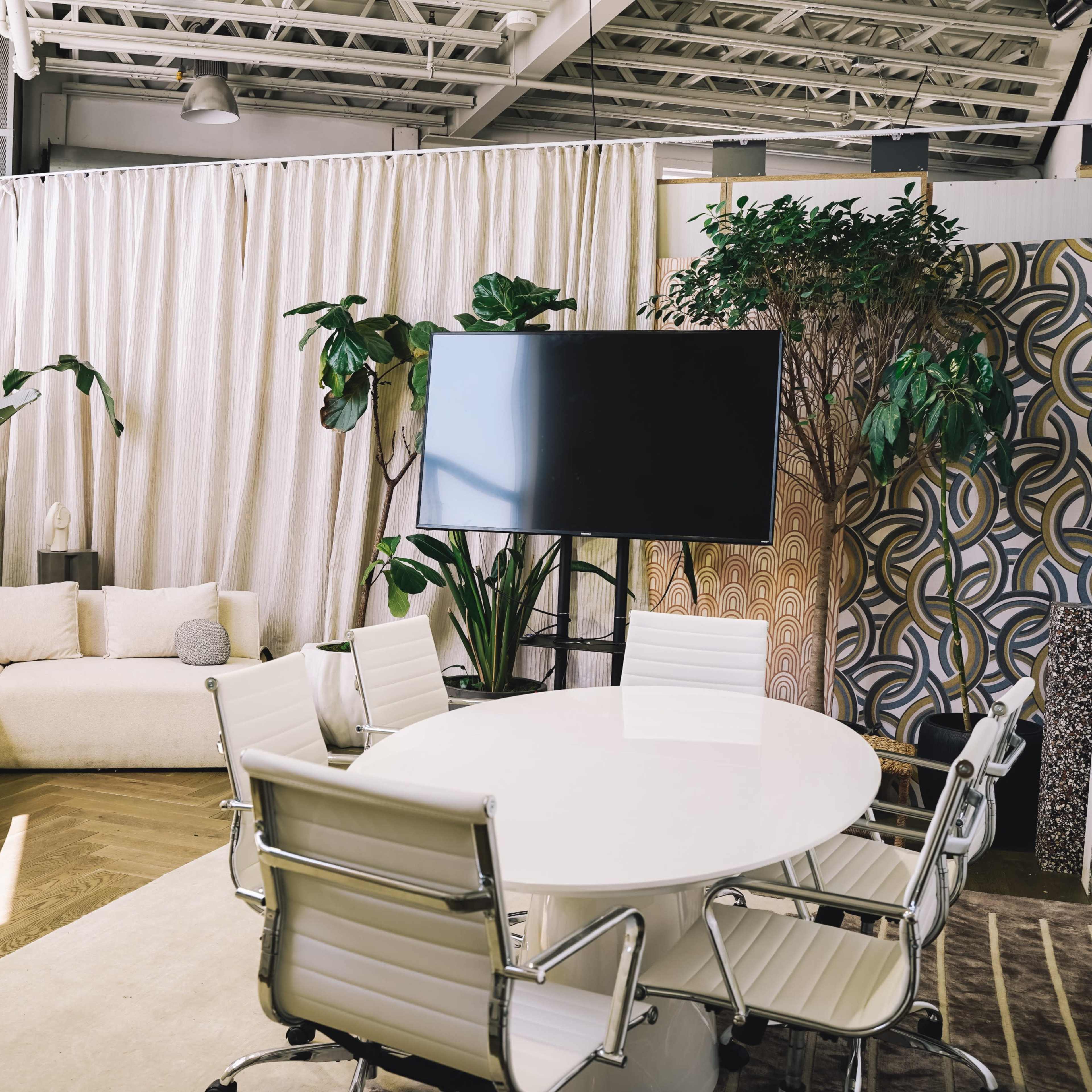 A modern meeting room with a round white table, swivel chairs, a large screen, and decorative plants set against a backdrop of curtains and patterned walls.
