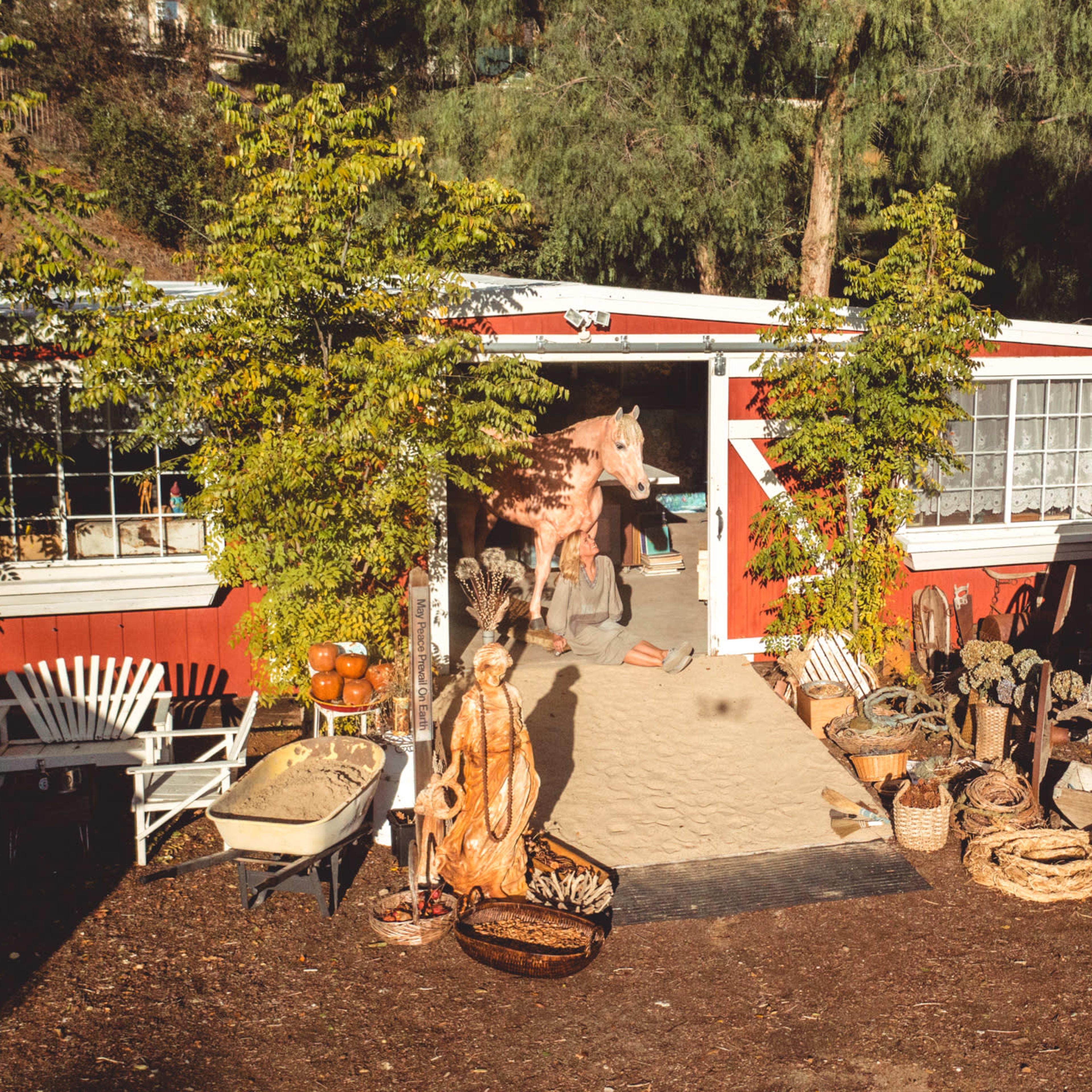 A red building with white trim features an entrance surrounded by various plants and rustic decor, including baskets, a wooden cart, and an array of sculptures.