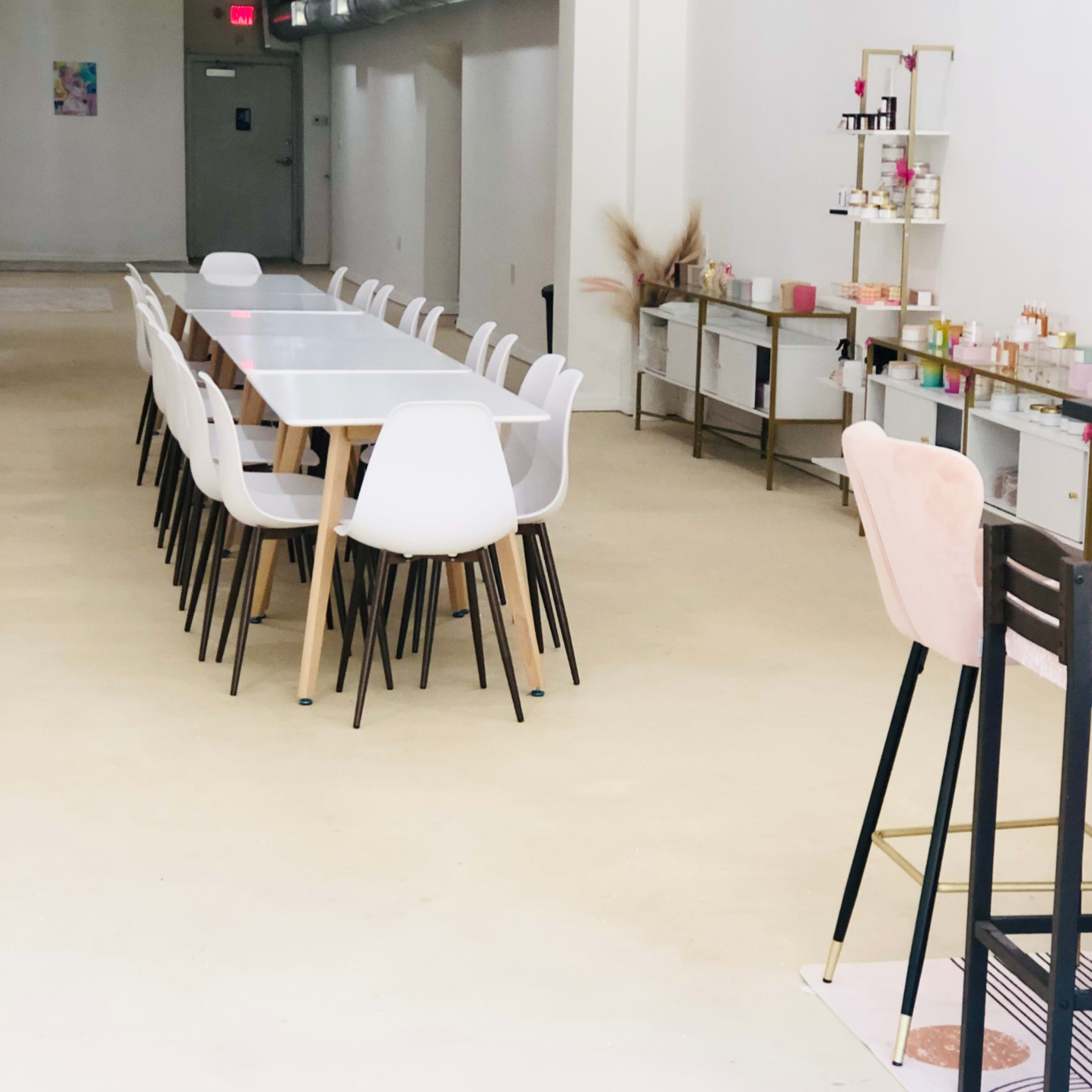 A long table with white chairs is set up in a spacious, minimalistic room featuring storage units and a bar area along one side.