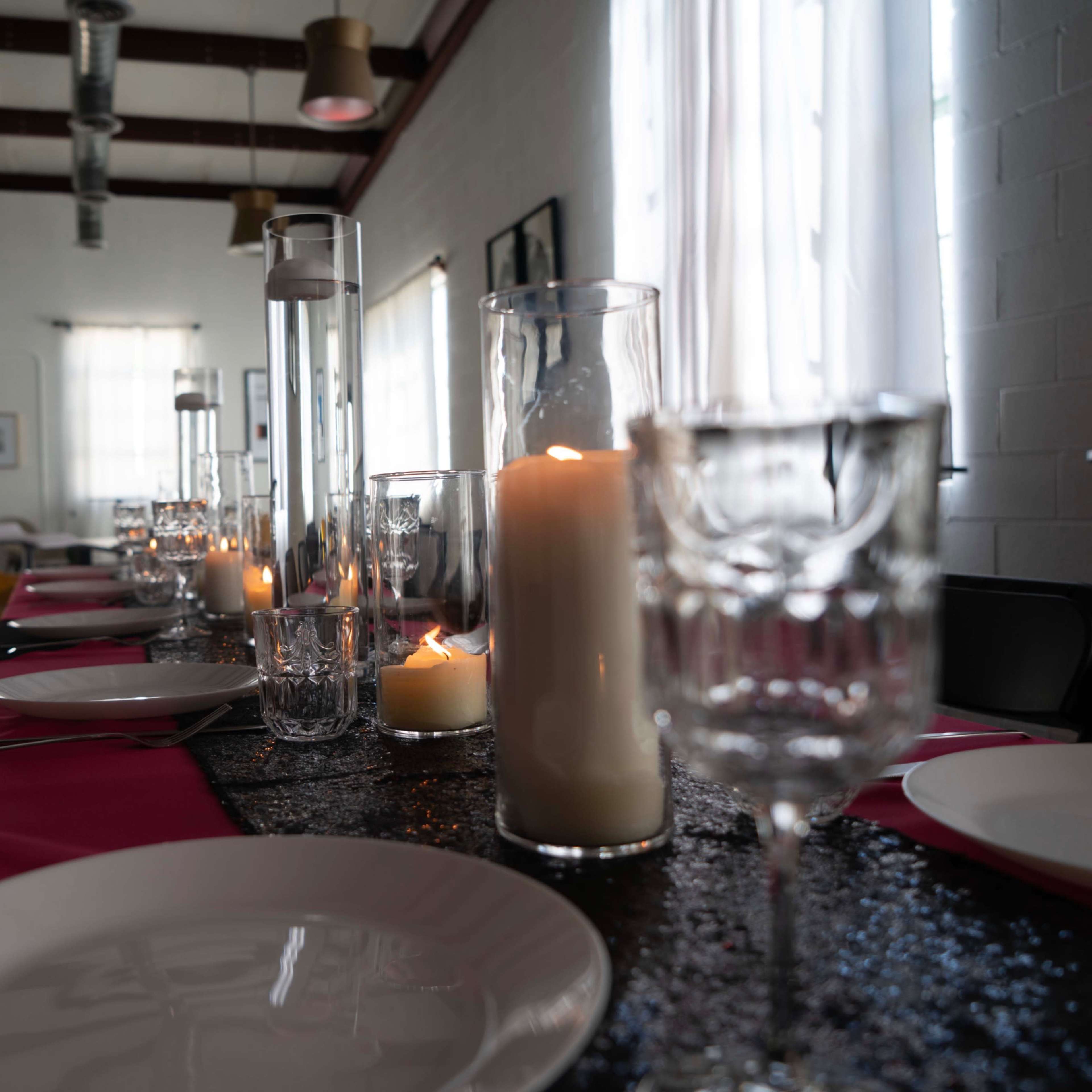 A long dining table is set with white plates, crystal glasses, and decorative candles, illuminated by natural light coming through large windows.