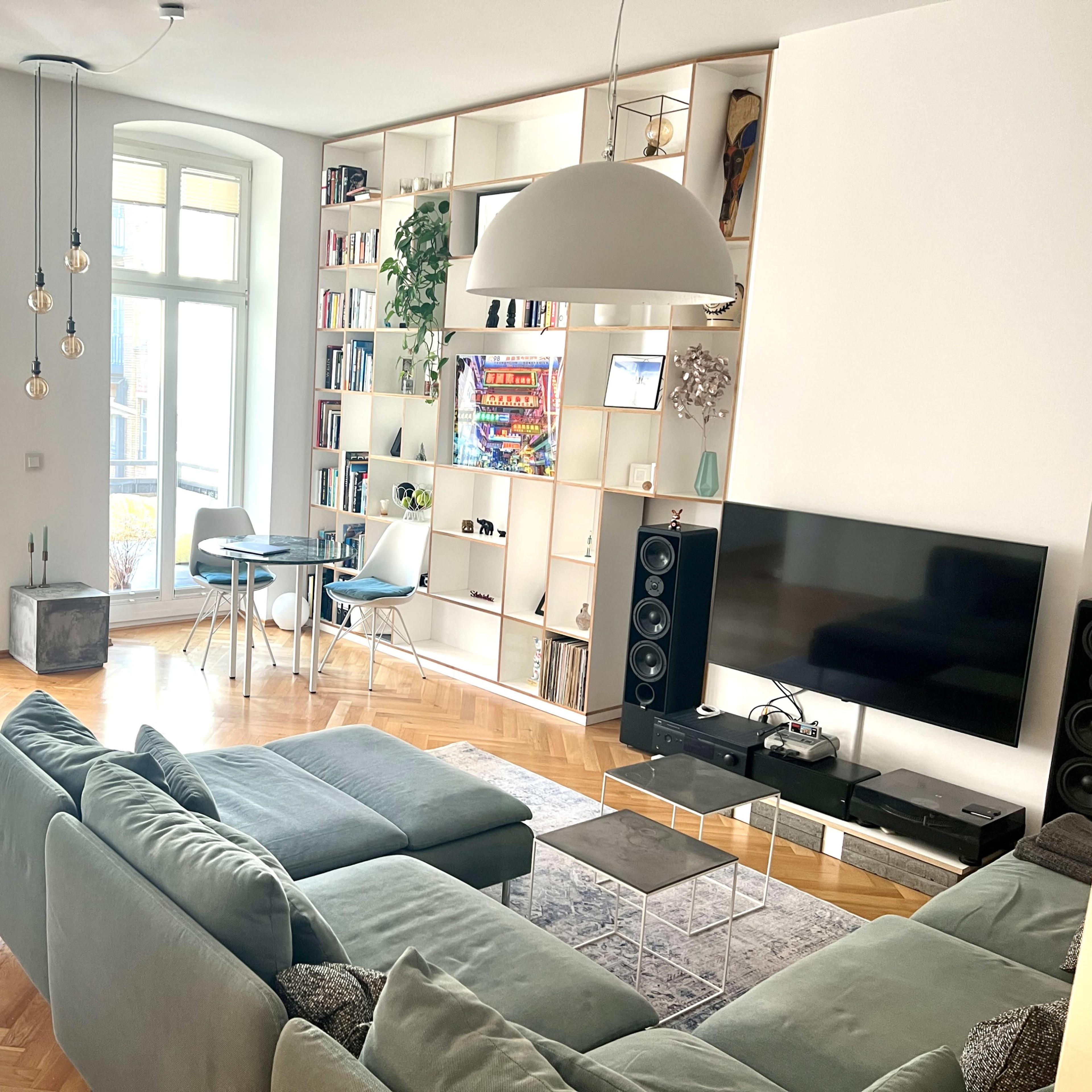 A modern living room featuring a light green sectional sofa, a glass coffee table, a television mounted on the wall, and a bookshelf filled with books, all with large windows allowing natural light.