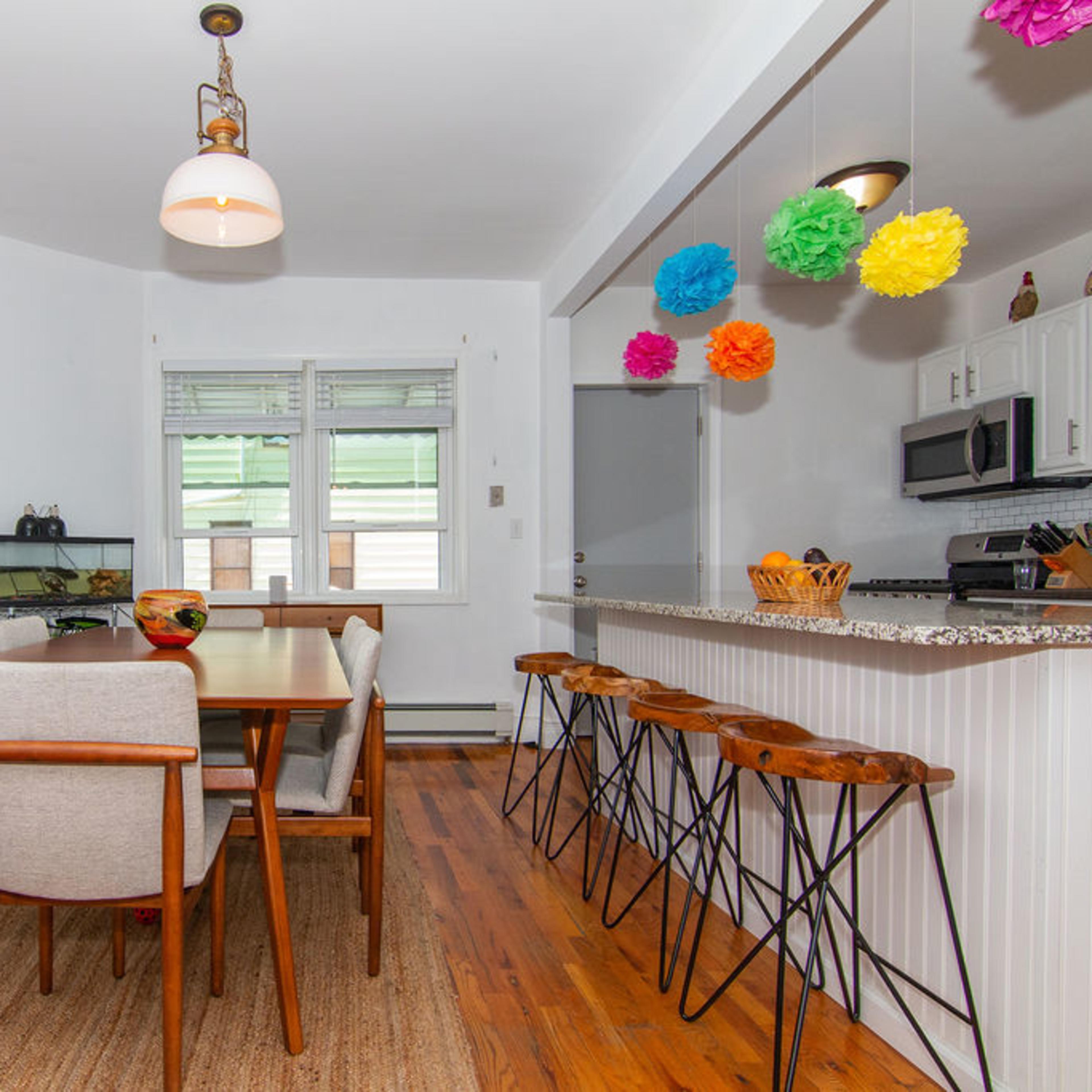 A modern dining area features a wooden table surrounded by six chairs, with a colorful artwork on the wall and a kitchen counter with bar stools in the background.