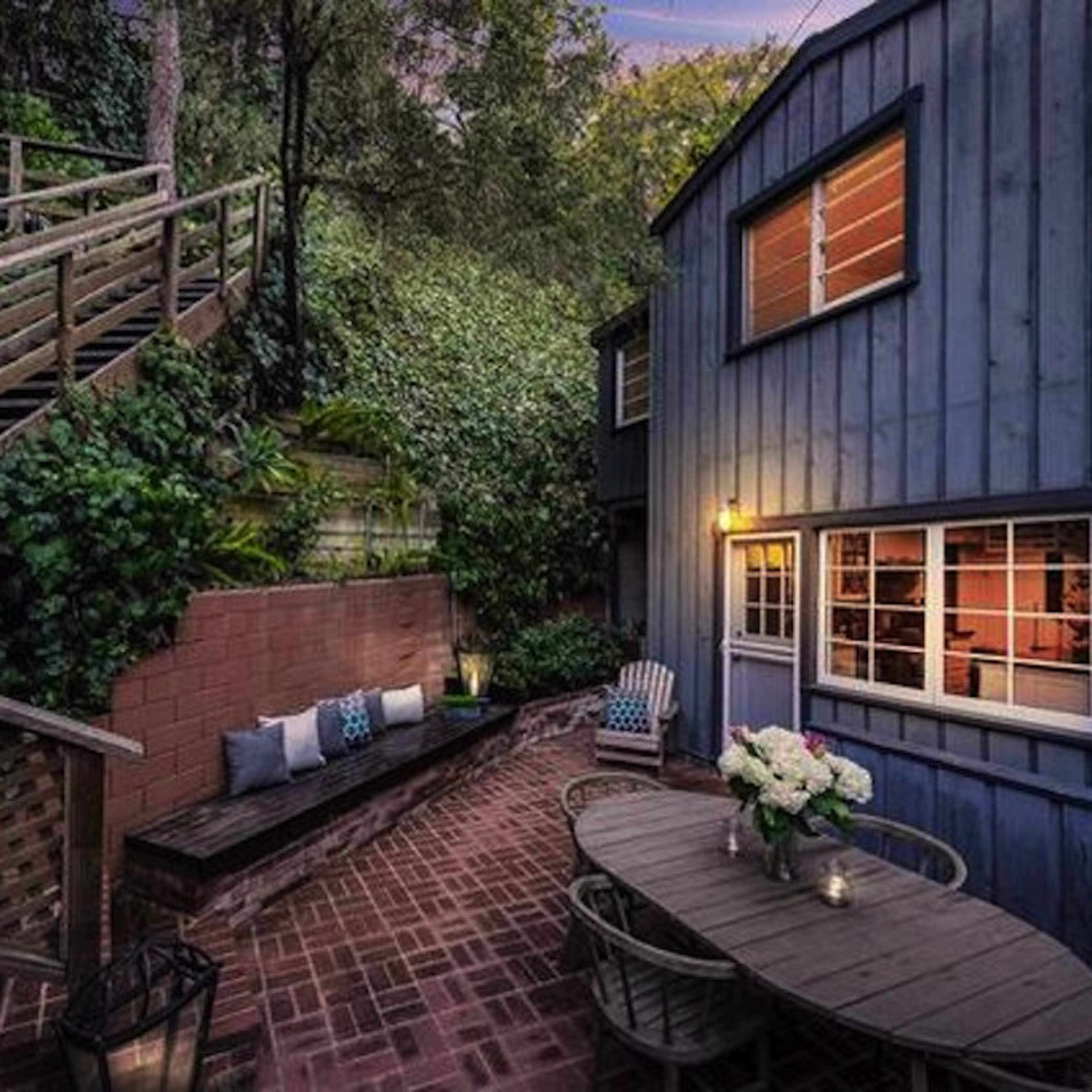 A blue wooden house is nestled among greenery, featuring a brick patio with outdoor seating and a staircase leading up to a higher level.