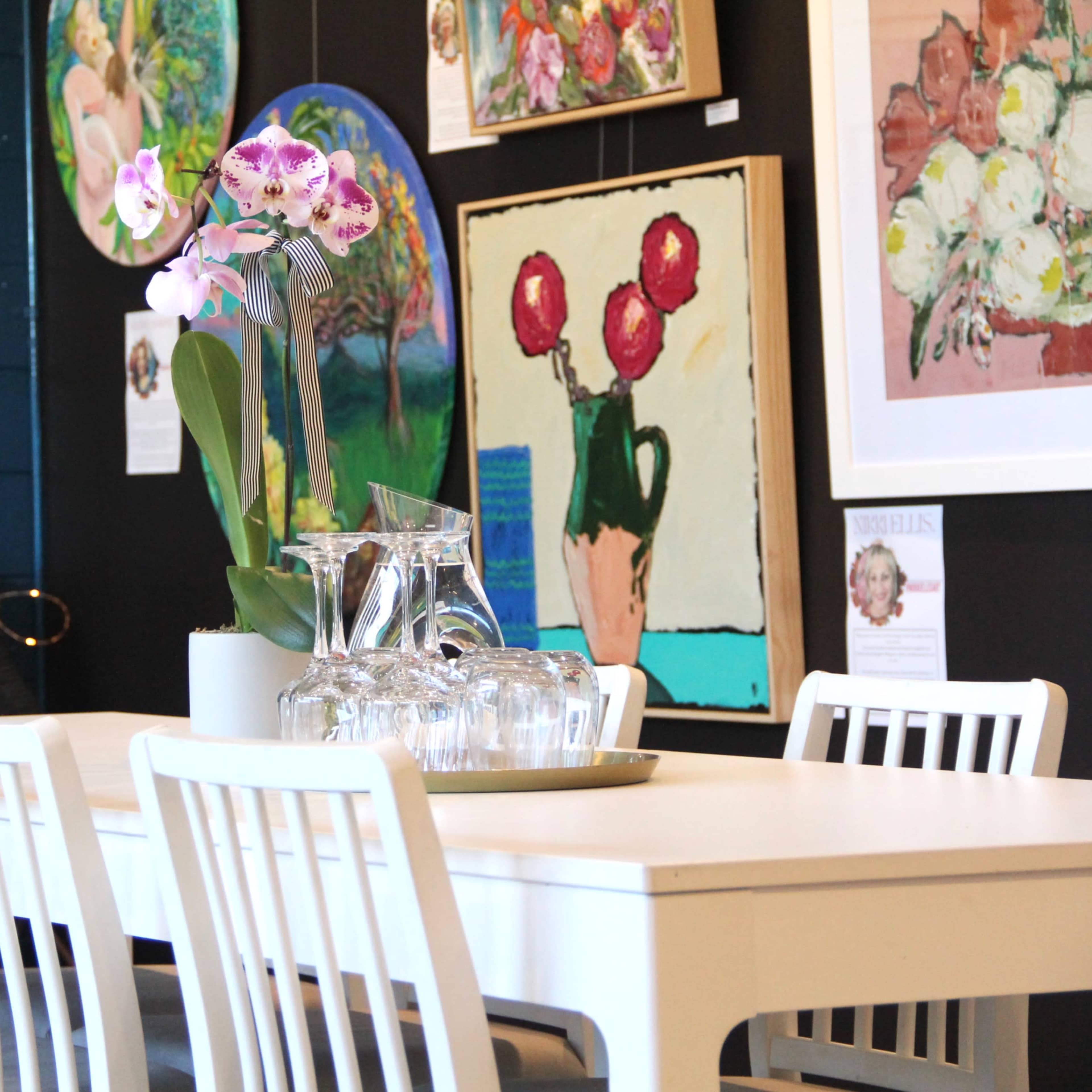 A white table is set with a pitcher and glasses, surrounded by chairs, in a room displaying colorful artwork on the walls.