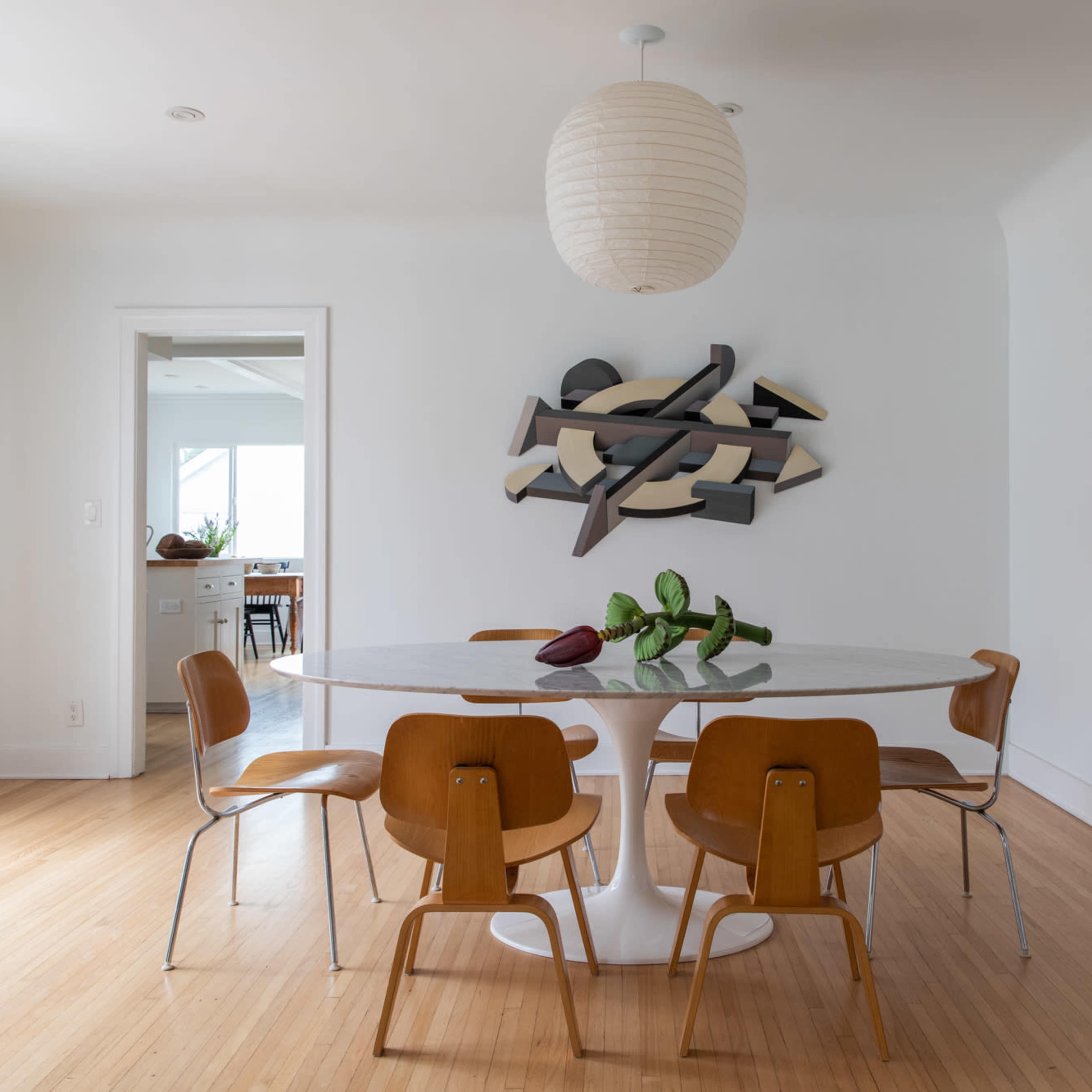 A dining area features a round table surrounded by four wooden chairs, with a large artwork on the wall opposite a door leading outside.