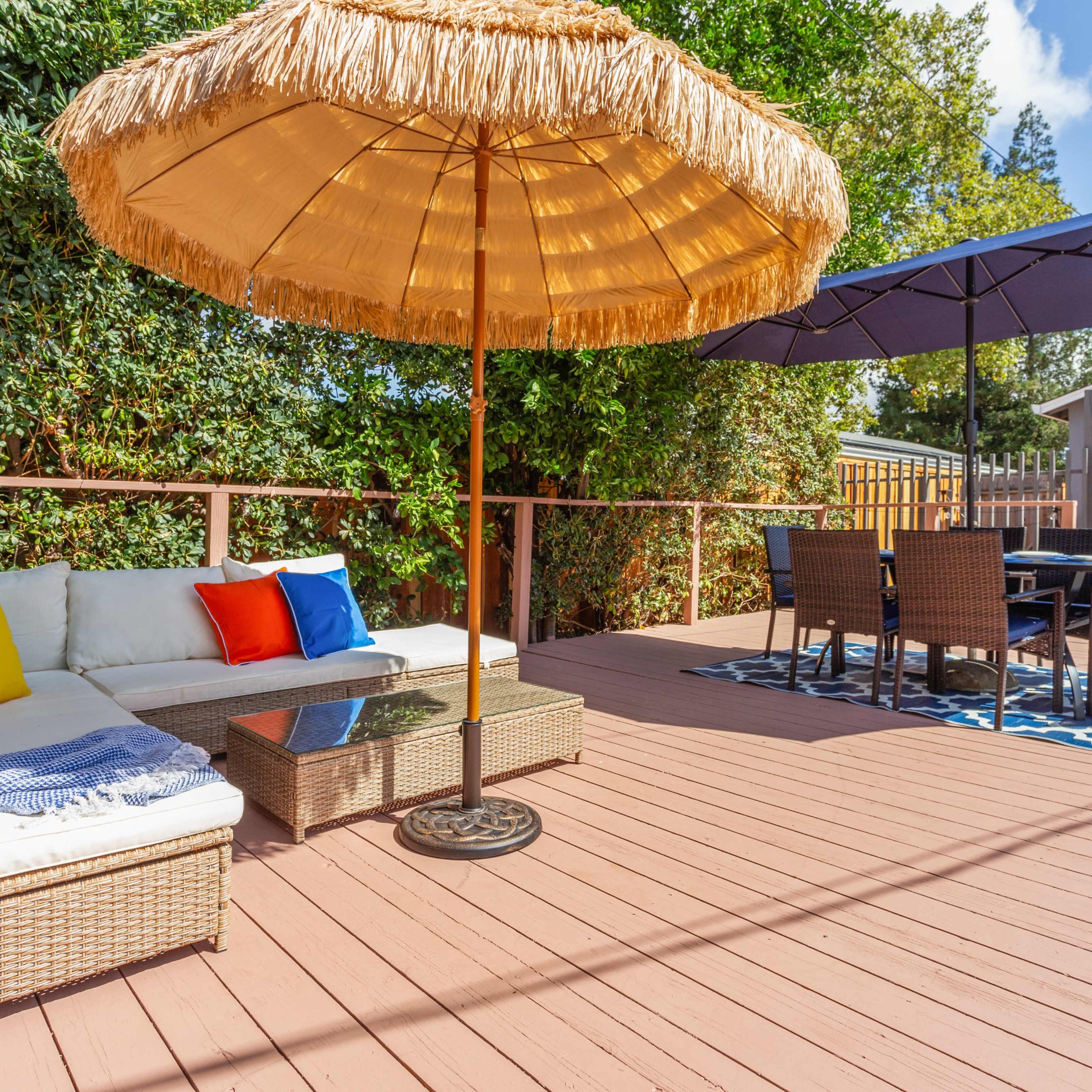 A patio features a wicker seating area with colorful pillows, a thatched umbrella, and a dining set arranged on a patterned rug.