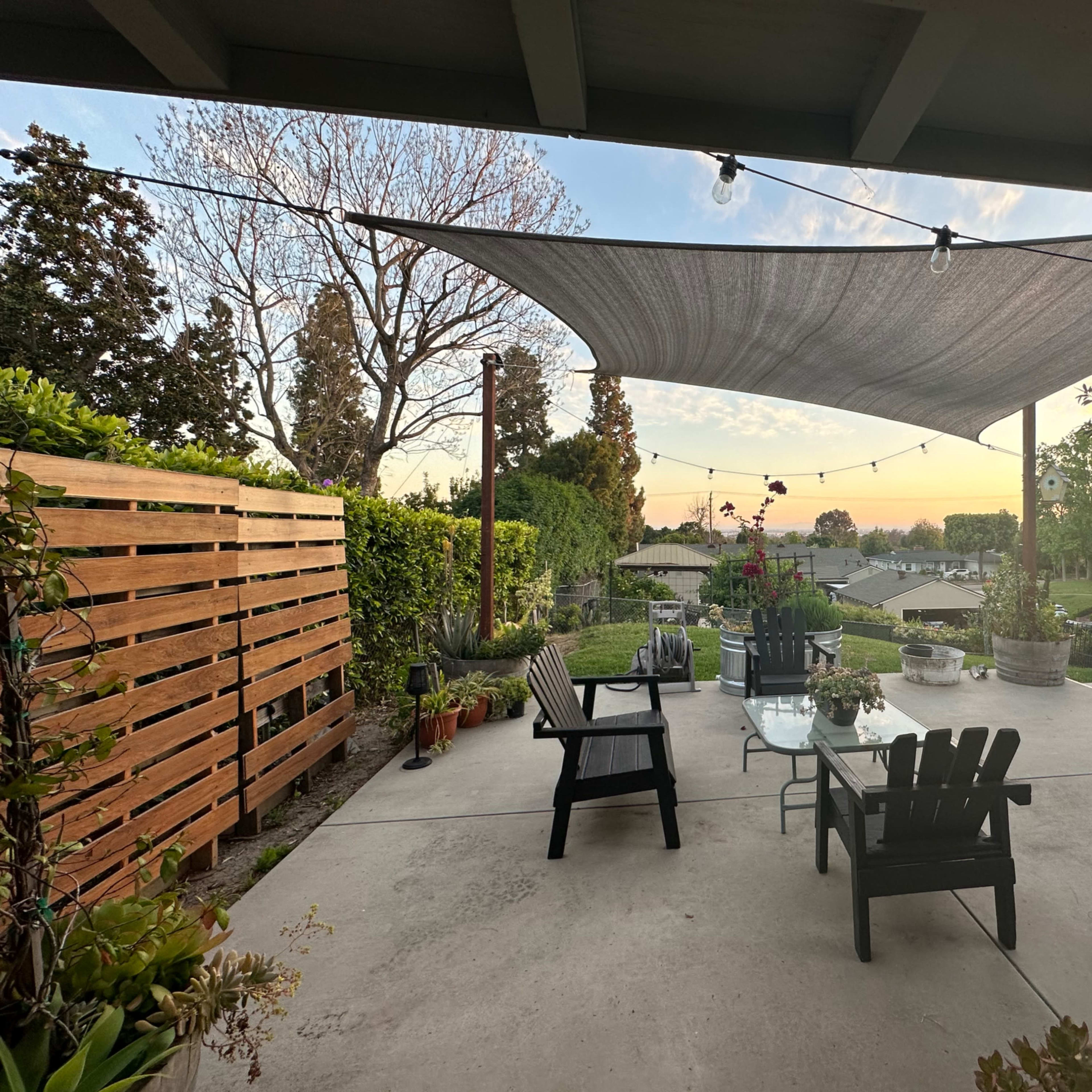 A patio with modern furniture, a shade sail overhead, and potted plants, overlooking a suburban area at sunset.
