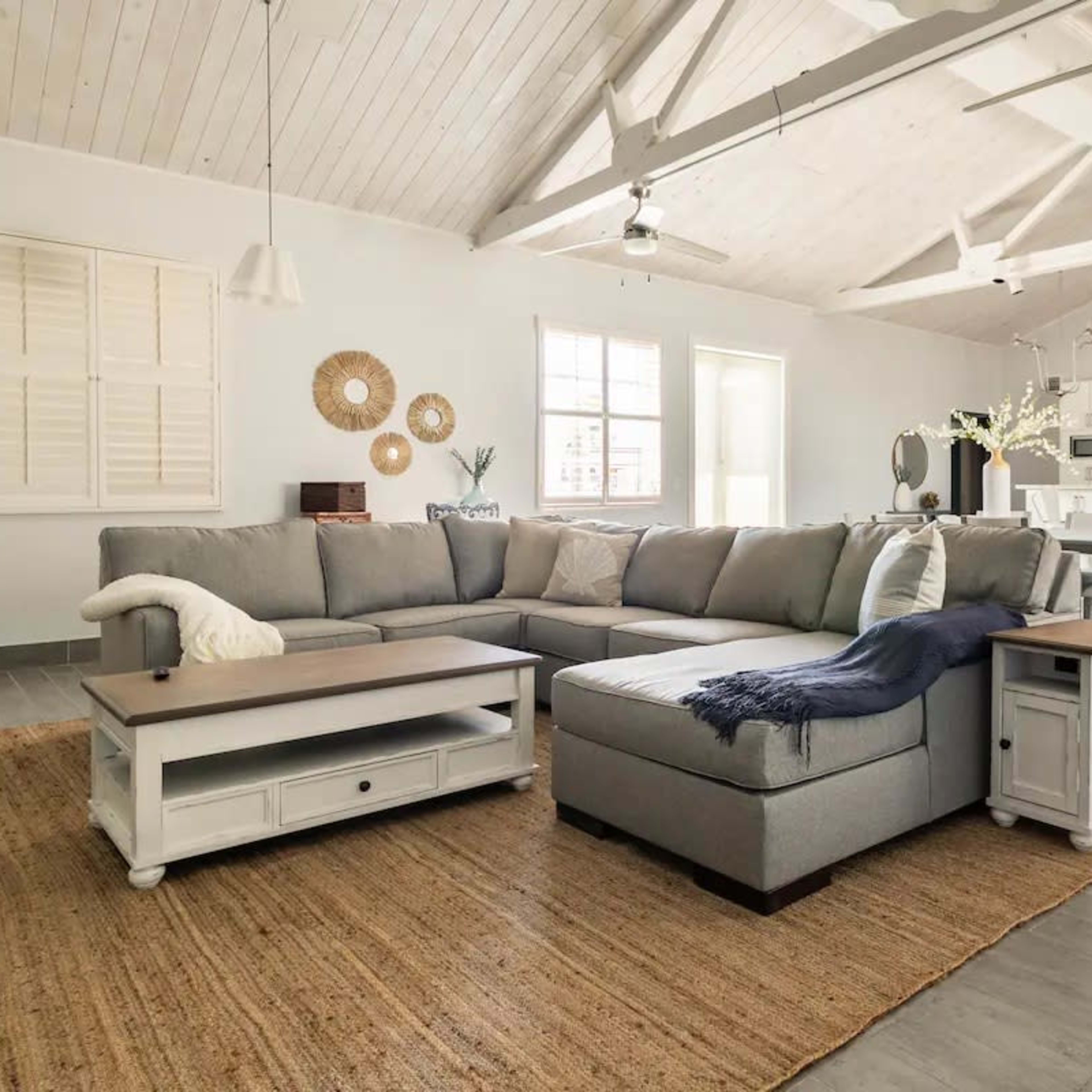 A spacious living room features a gray sectional sofa, a light wooden table, and a woven area rug beneath a vaulted ceiling.
