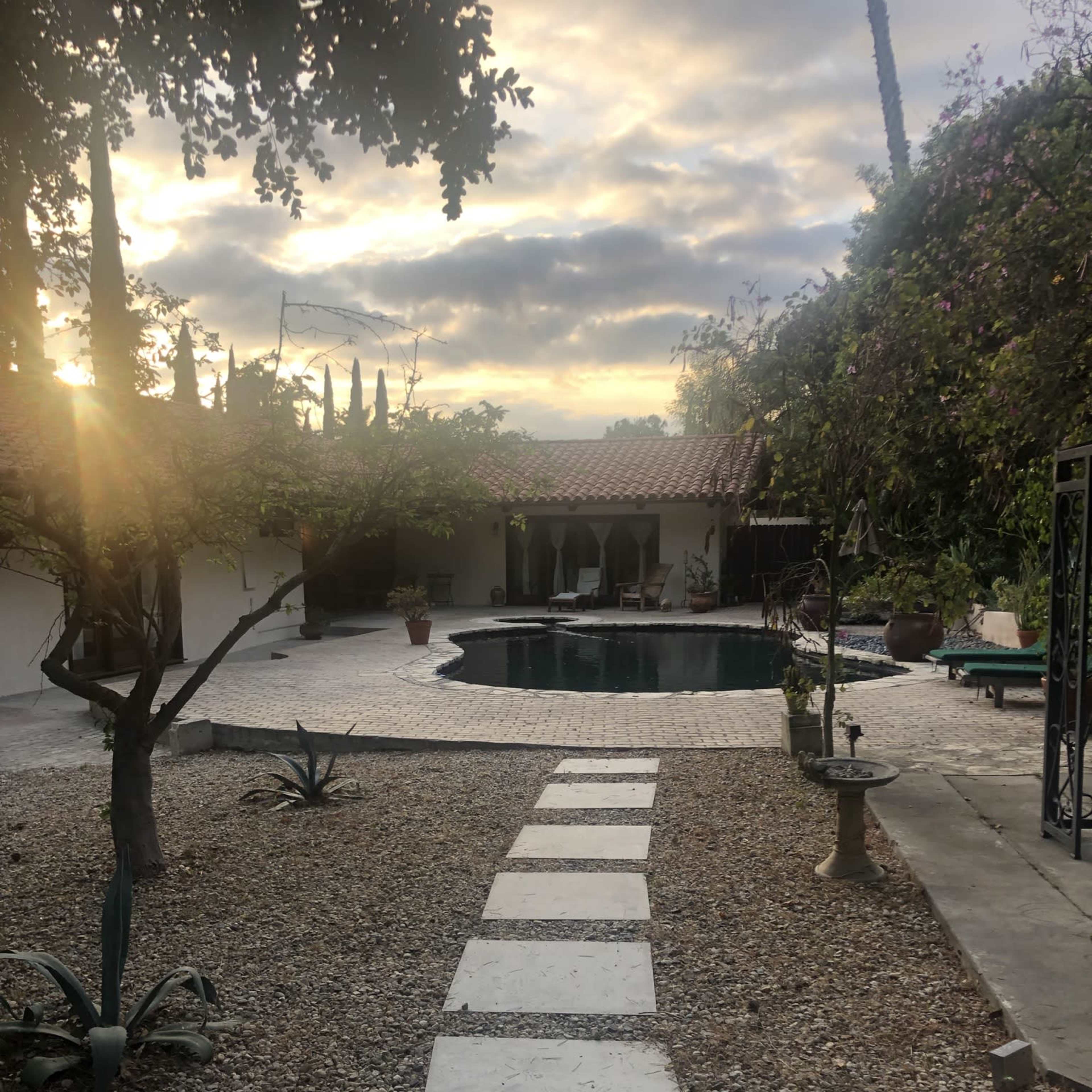 A stone pathway leads to a swimming pool surrounded by greenery and a patio under a cloudy sky.