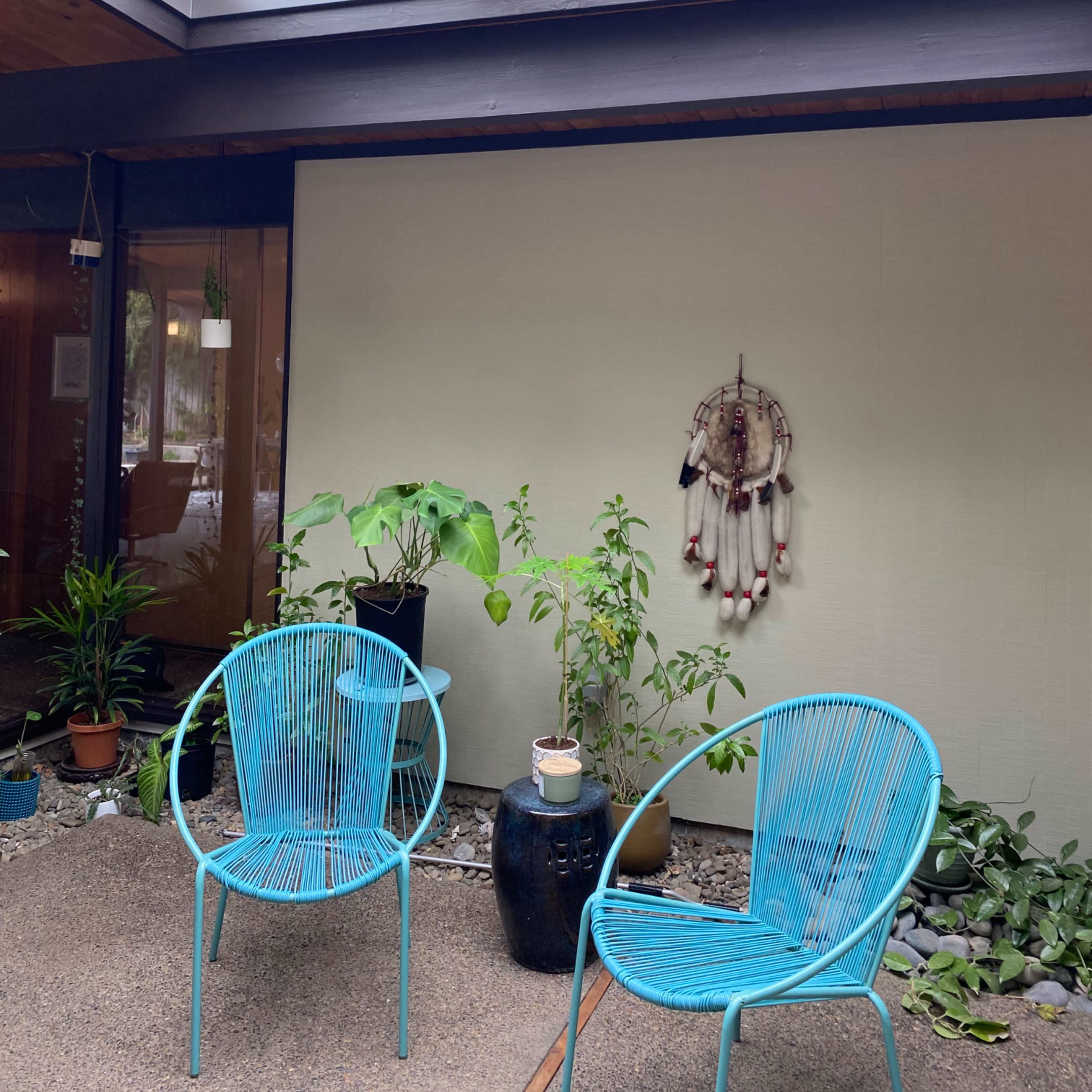 A modern patio area with two blue wire chairs, a potted plant, and a decorative dreamcatcher on the wall, all beneath a glass skylight.
