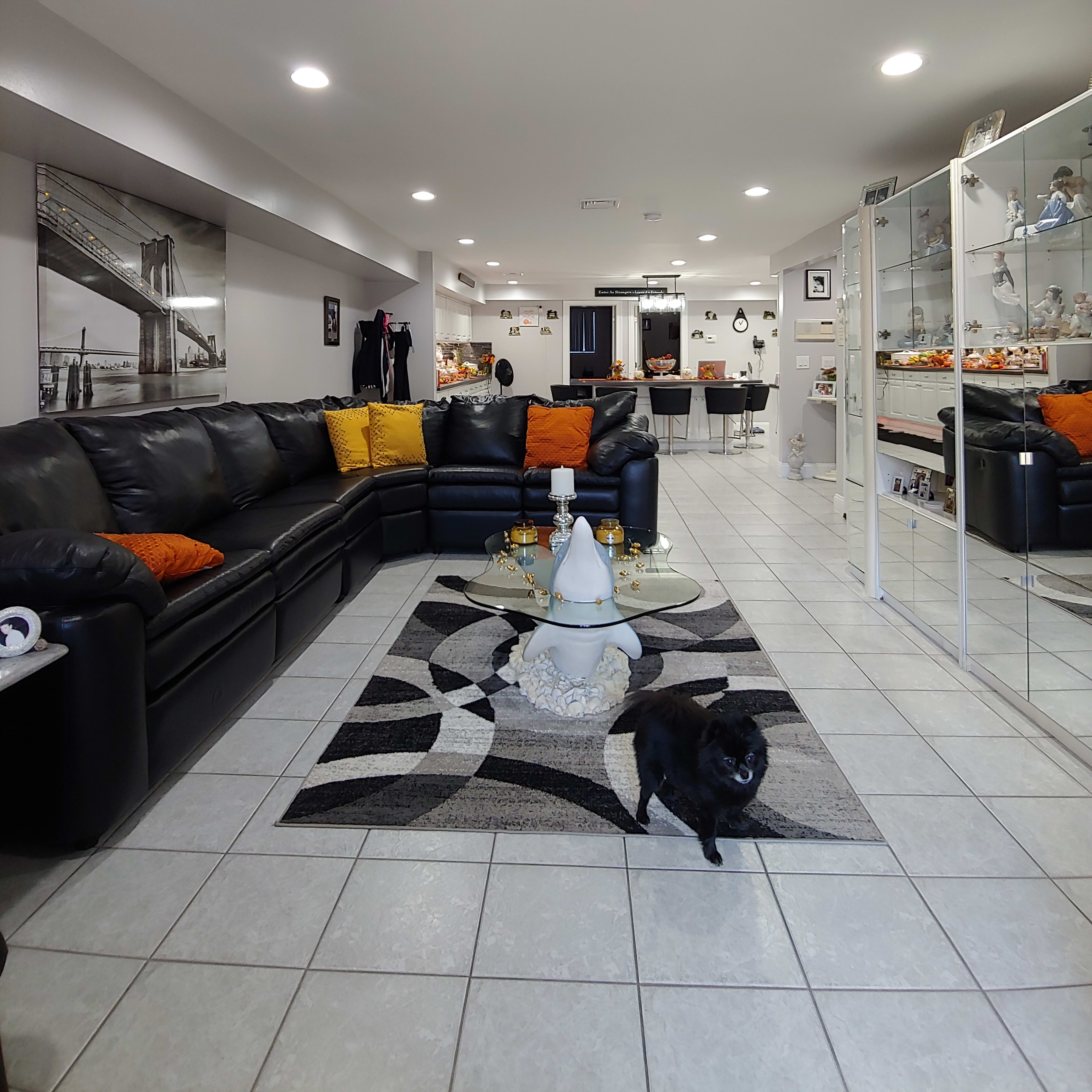 A modern basement living area with a black sectional couch, decorative pillows, a glass coffee table, and a small black dog on a tiled floor.