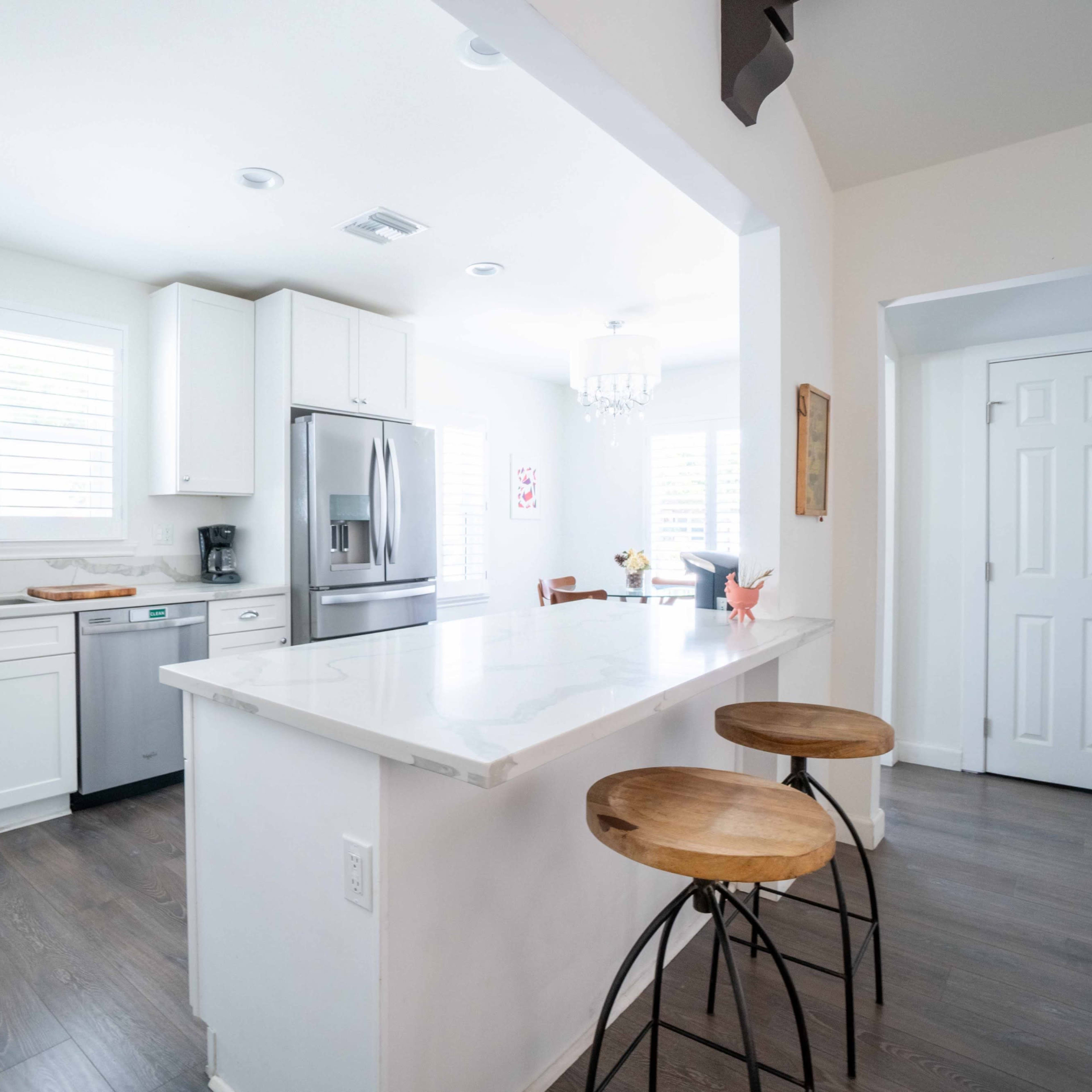 A modern kitchen featuring white cabinets, stainless steel appliances, and a countertop with two bar stools, adjacent to a dining area.