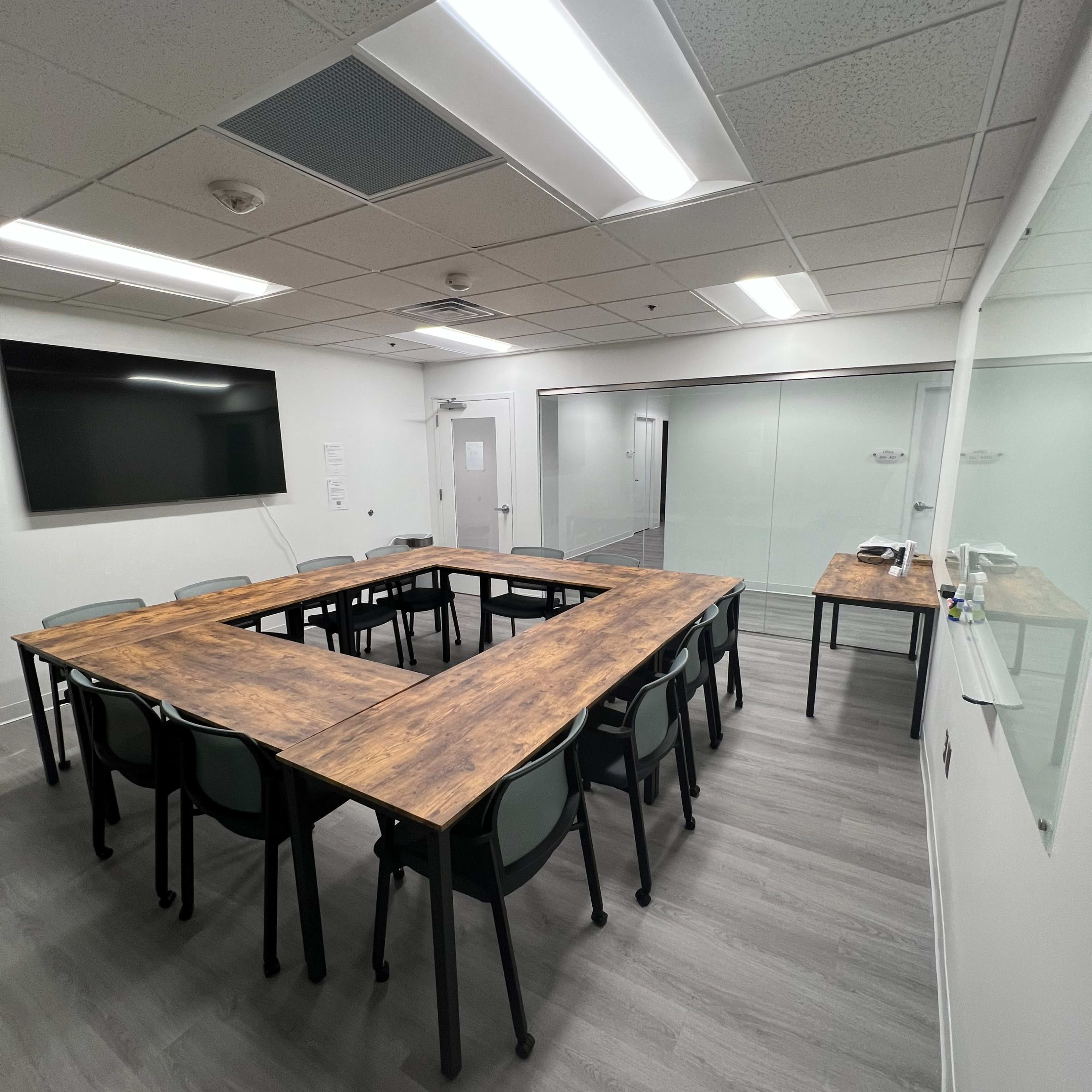 A conference room features a large wooden table arranged in a U-shape with chairs around it and a television monitor mounted on the wall.