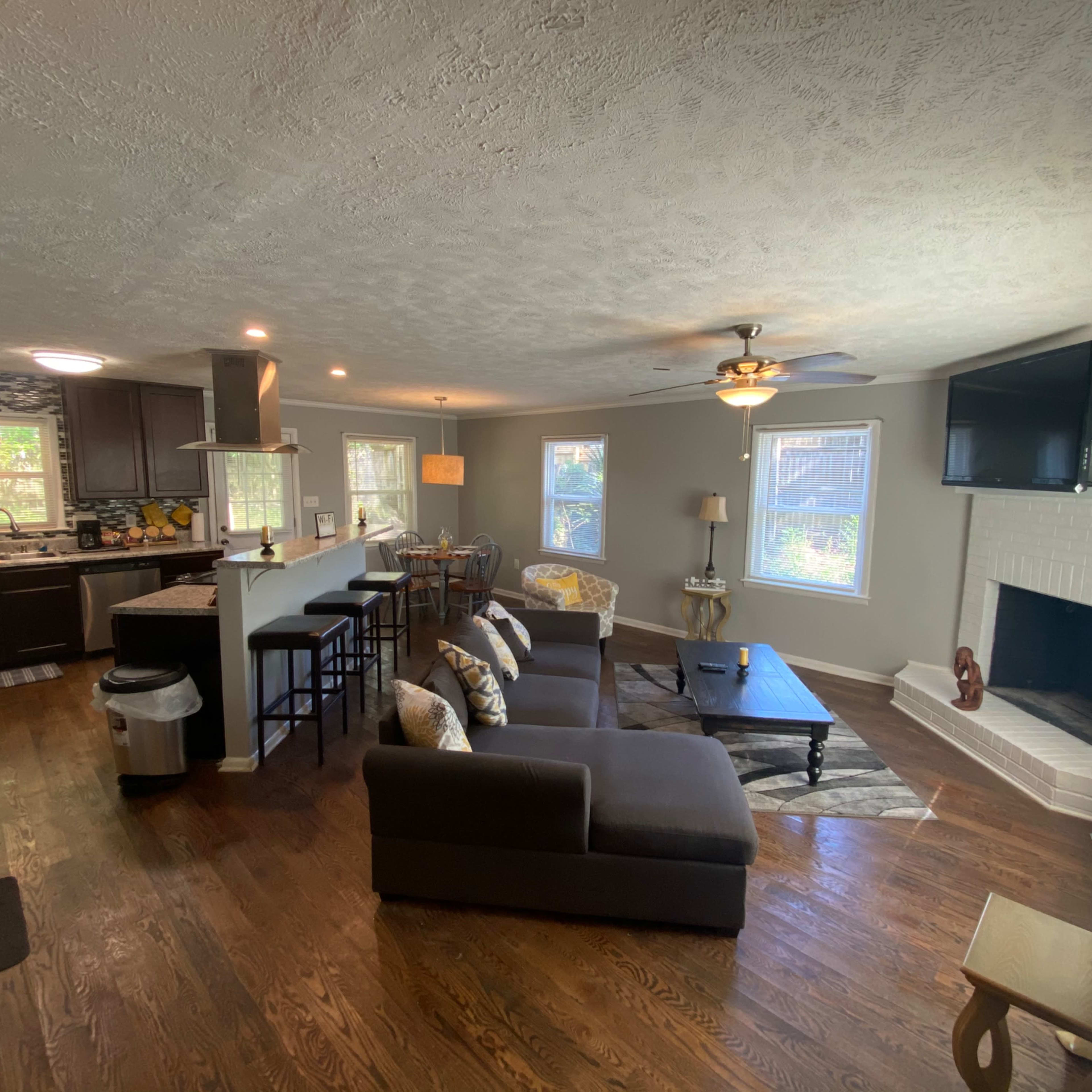 A modern living area with a dark sectional sofa, a dining space, and a kitchen visible in the background.