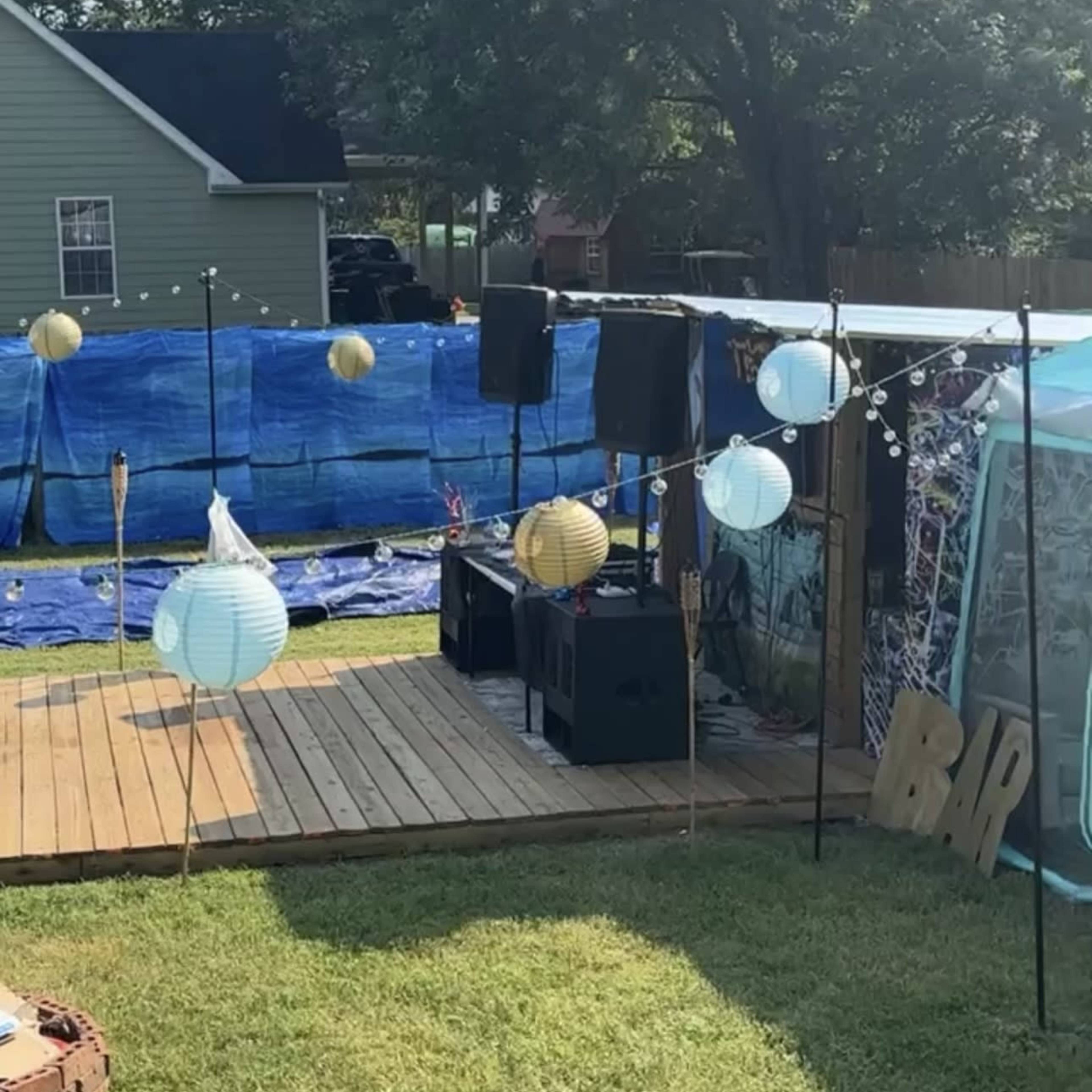 A backyard setup with a wooden deck, speakers, decorative lanterns, and a partially filled above-ground pool surrounded by a blue tarp.