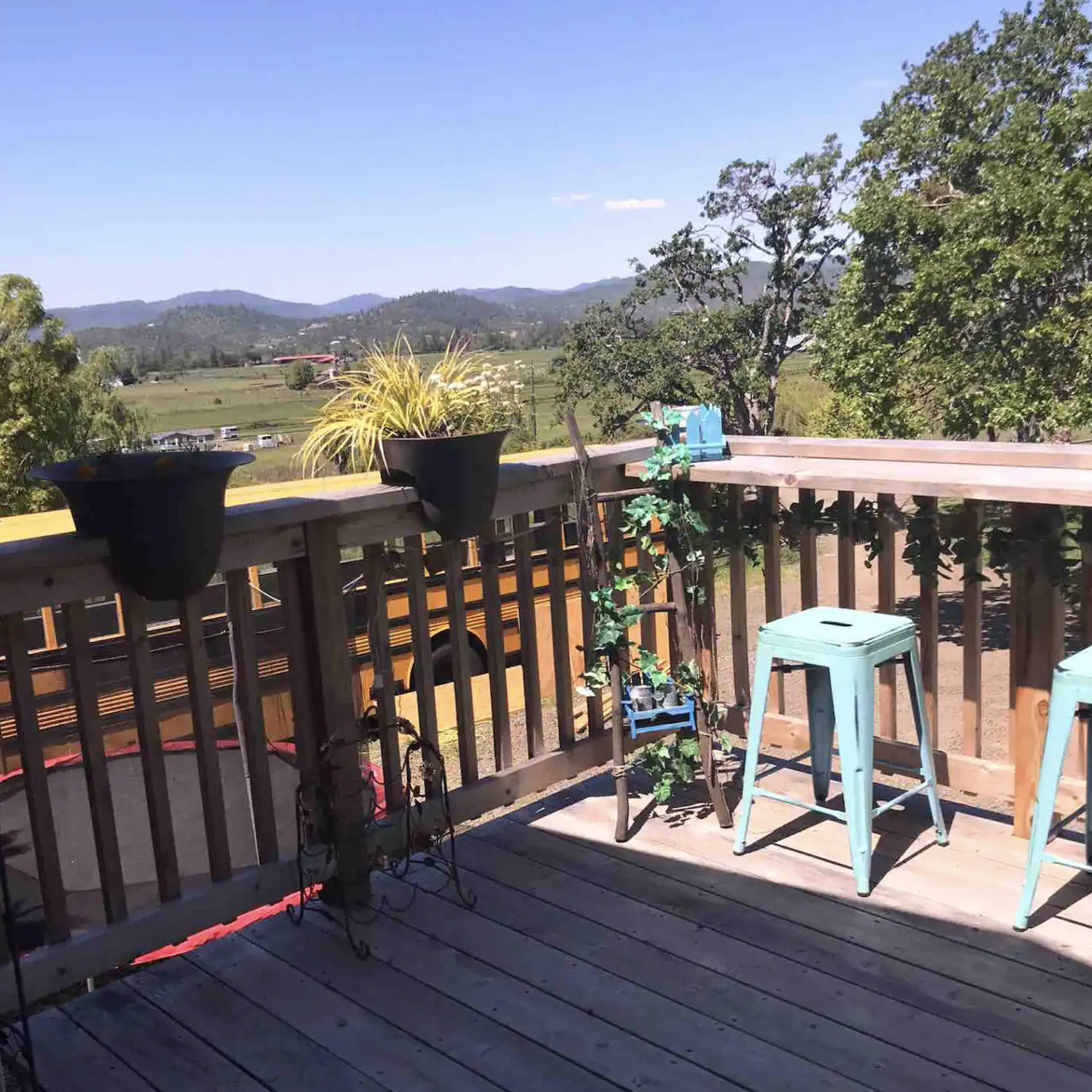 A wooden balcony features two blue stools, potted plants, and a view of a green landscape with mountains in the distance.