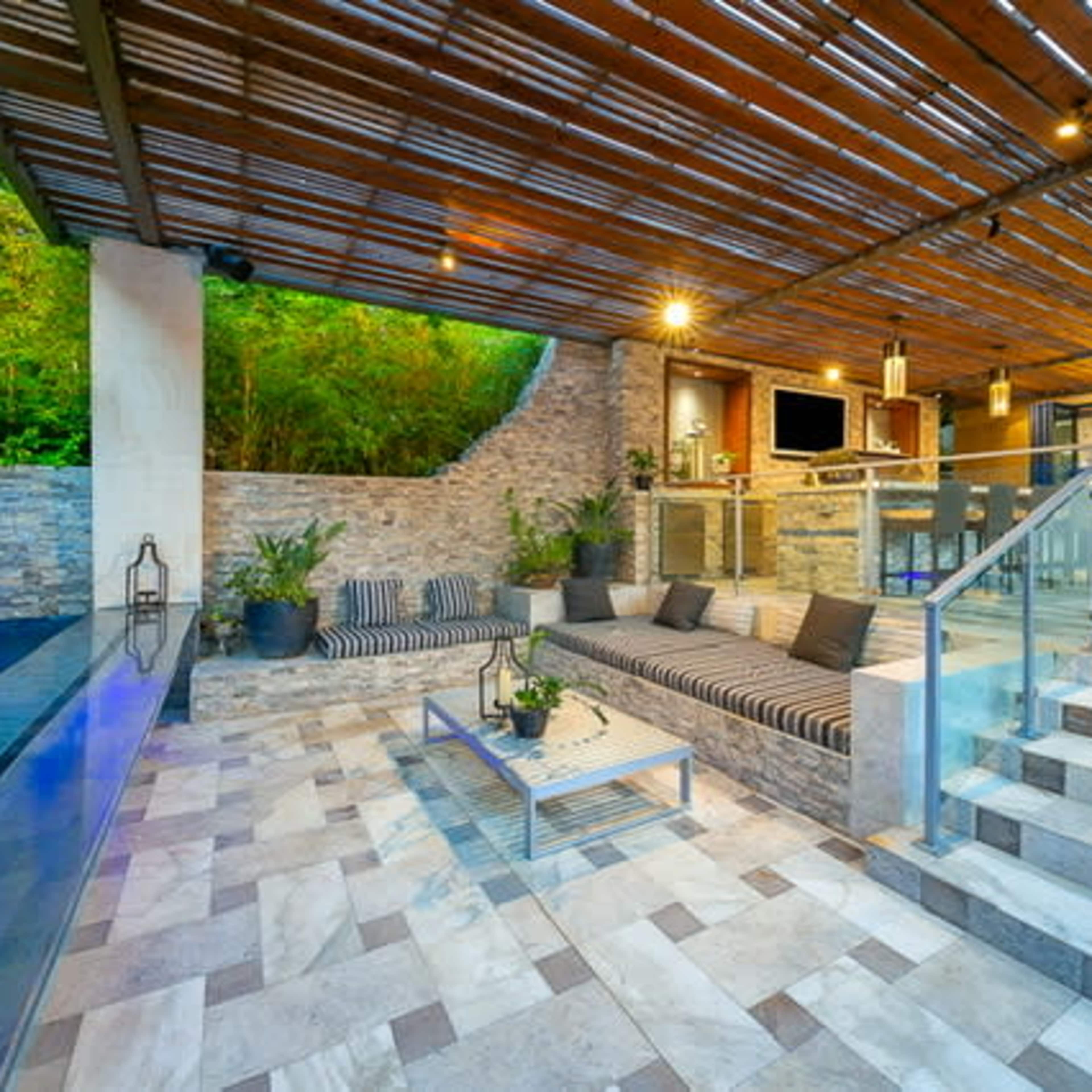 A modern outdoor living space with a lounge area, a stone wall, a swimming pool, and stairs leading to an indoor kitchen.