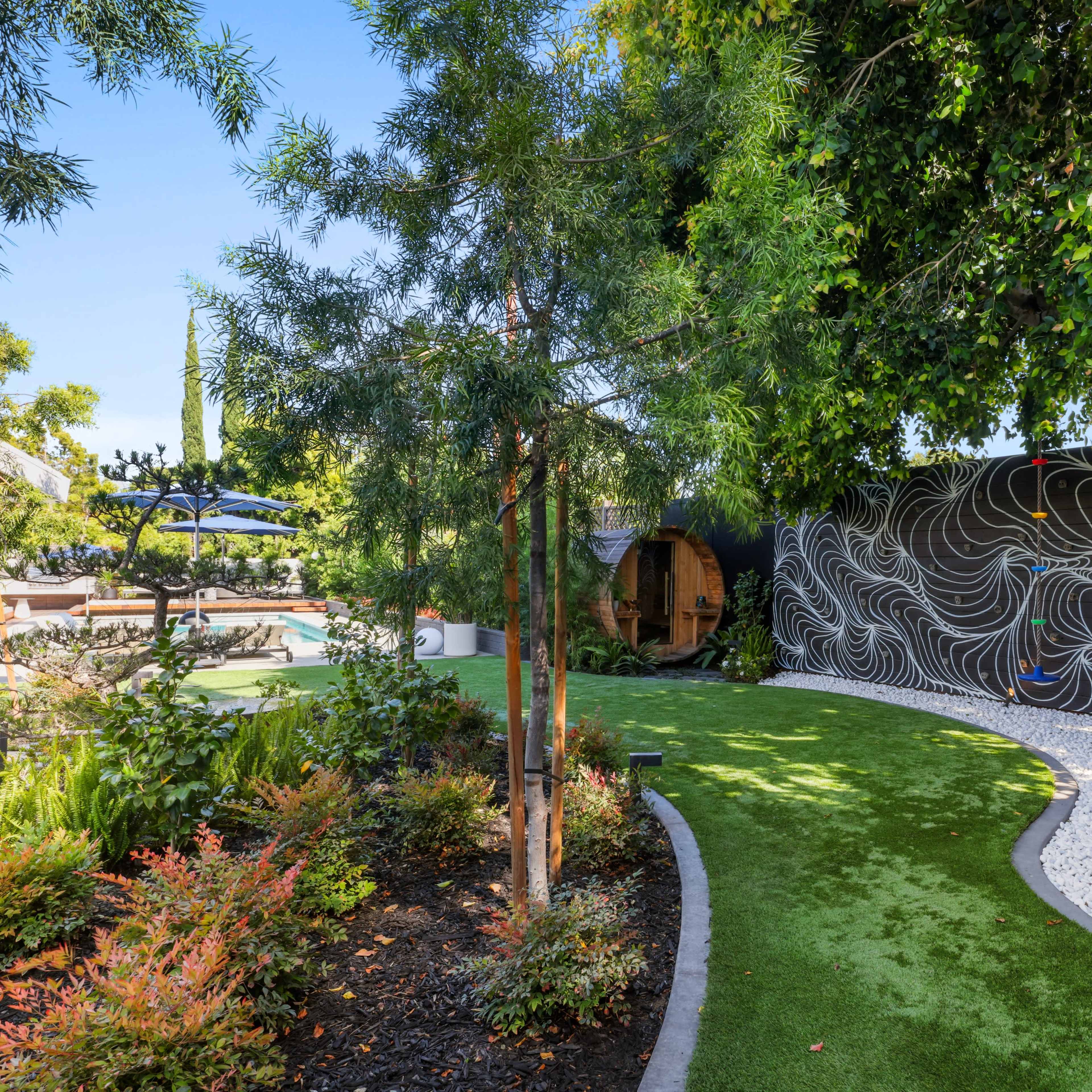 A landscaped backyard features a winding path of artificial grass bordered by shrubs and decorative rocks, leading to a seating area with a hanging chair and a pool in the background.