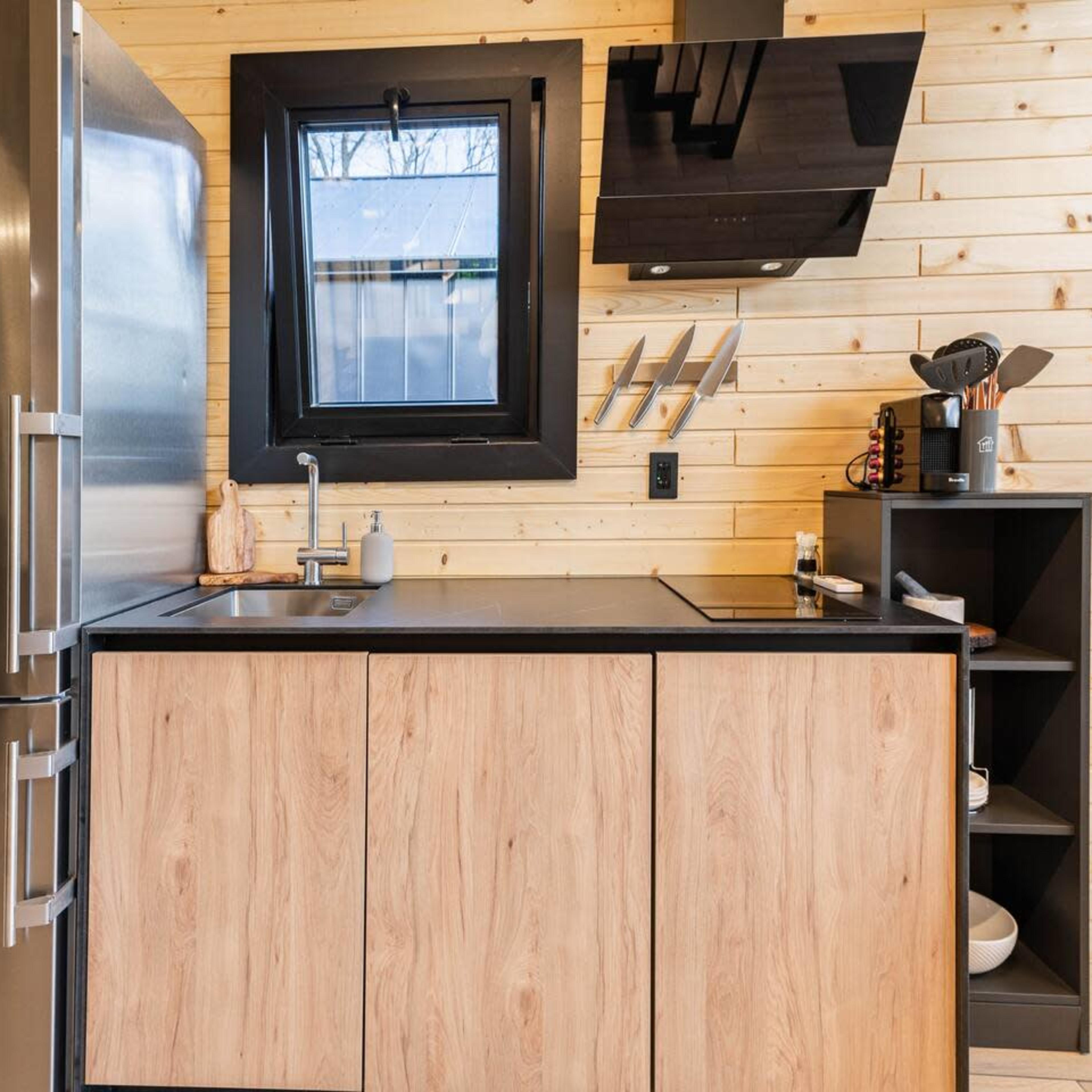 A modern kitchen with wooden walls, a stainless steel refrigerator, a black countertop, and open shelving for kitchen utensils.