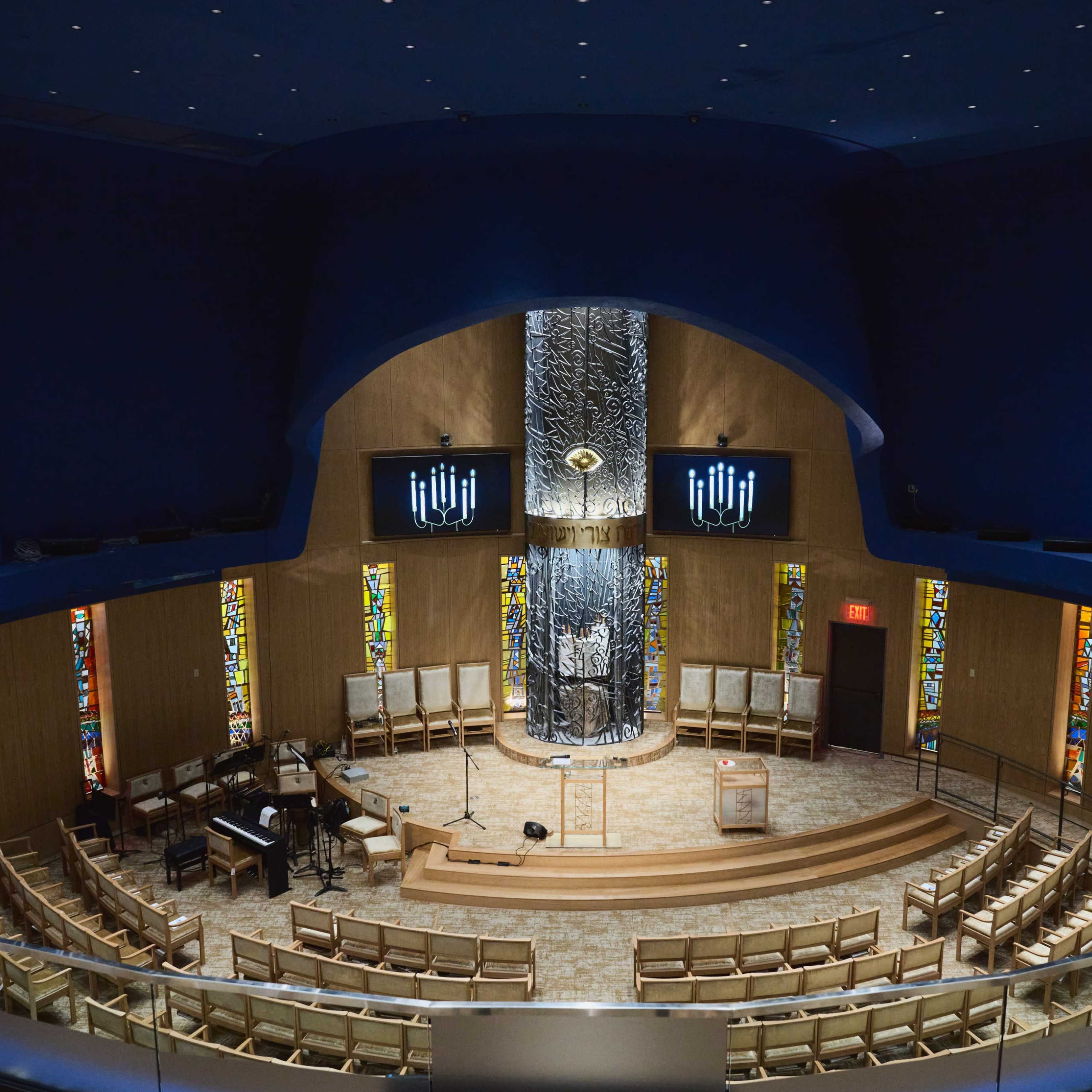 A modern interior of a circular synagogue featuring a central podium, stained glass windows, and seating arranged in multiple rows.