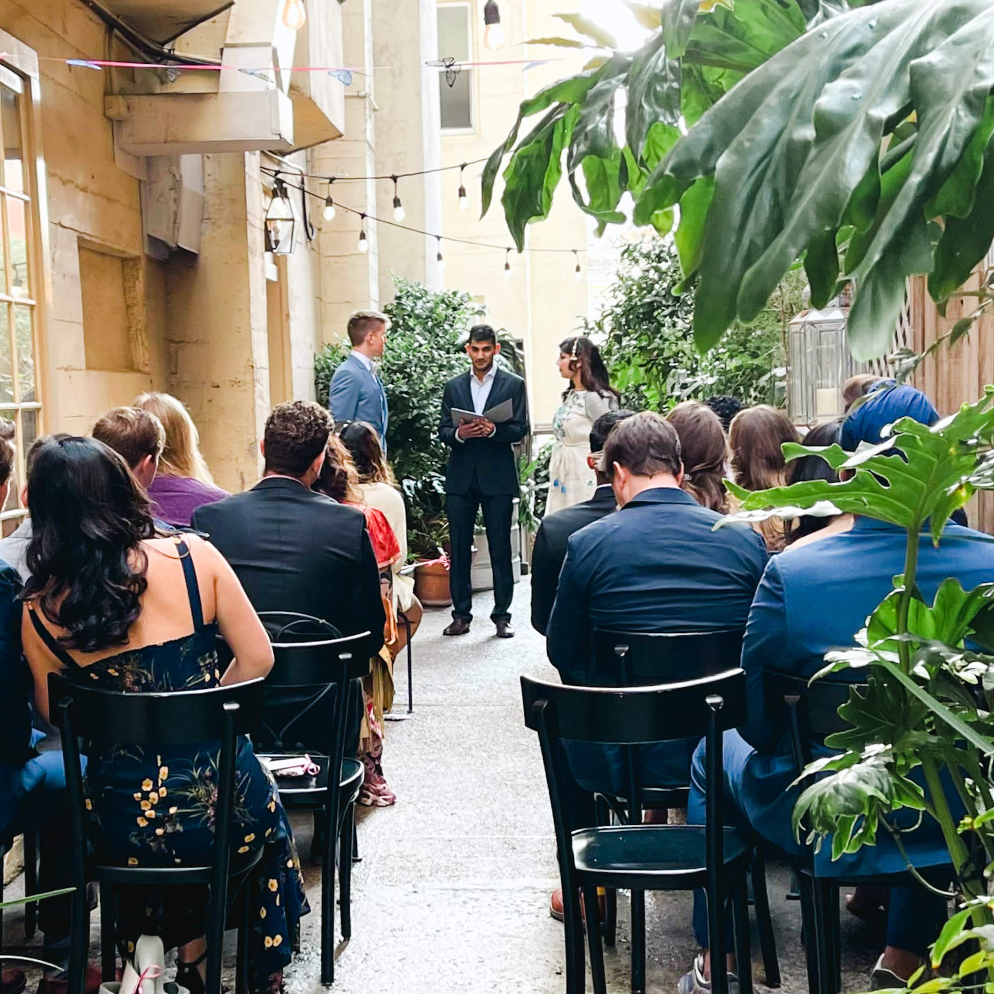 A couple stands at the front of an outdoor venue, surrounded by guests seated in chairs, while an officiant appears to be conducting a ceremony.