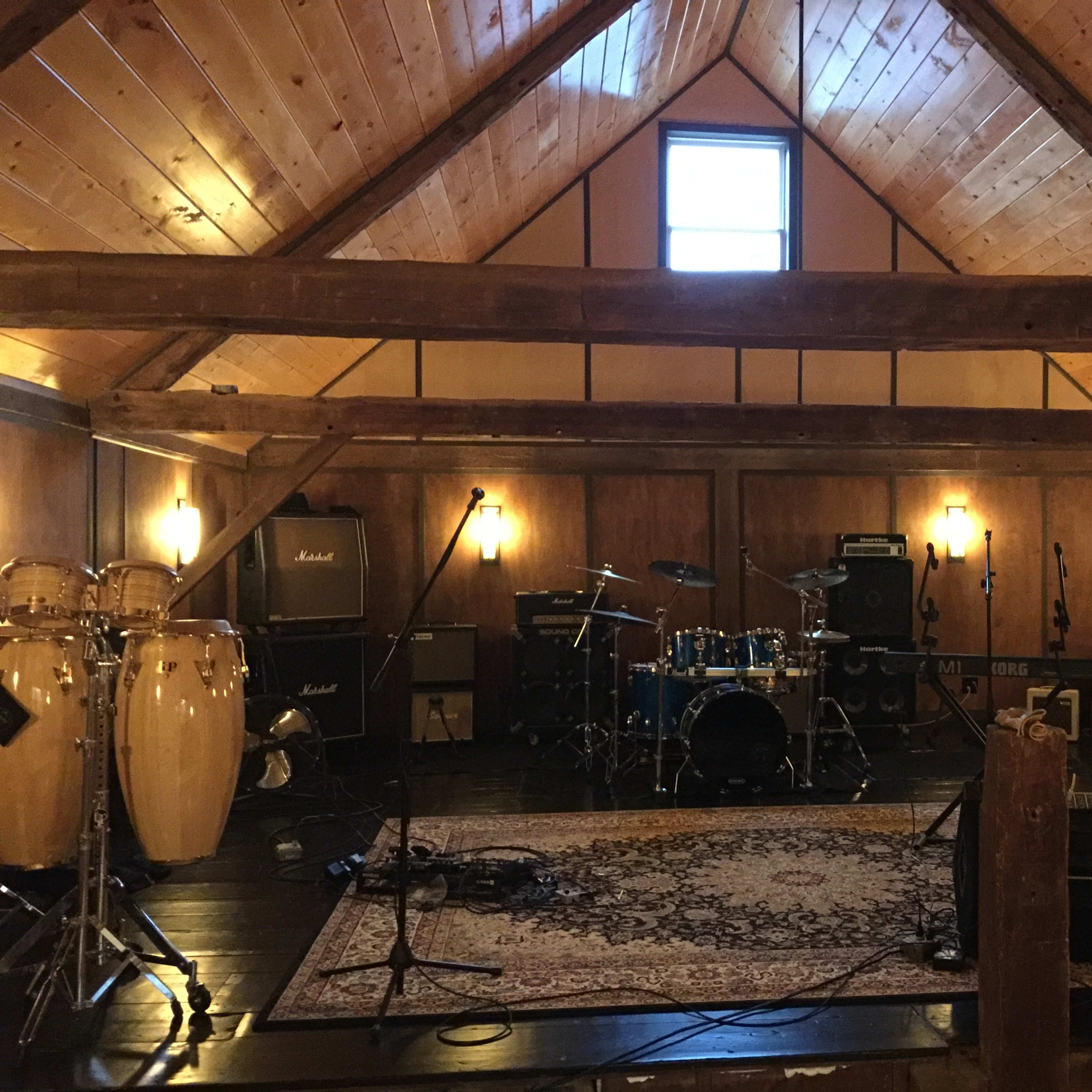 A music studio with various instruments, including drums and congas, set up in a wooden room with high ceilings and exposed beams.