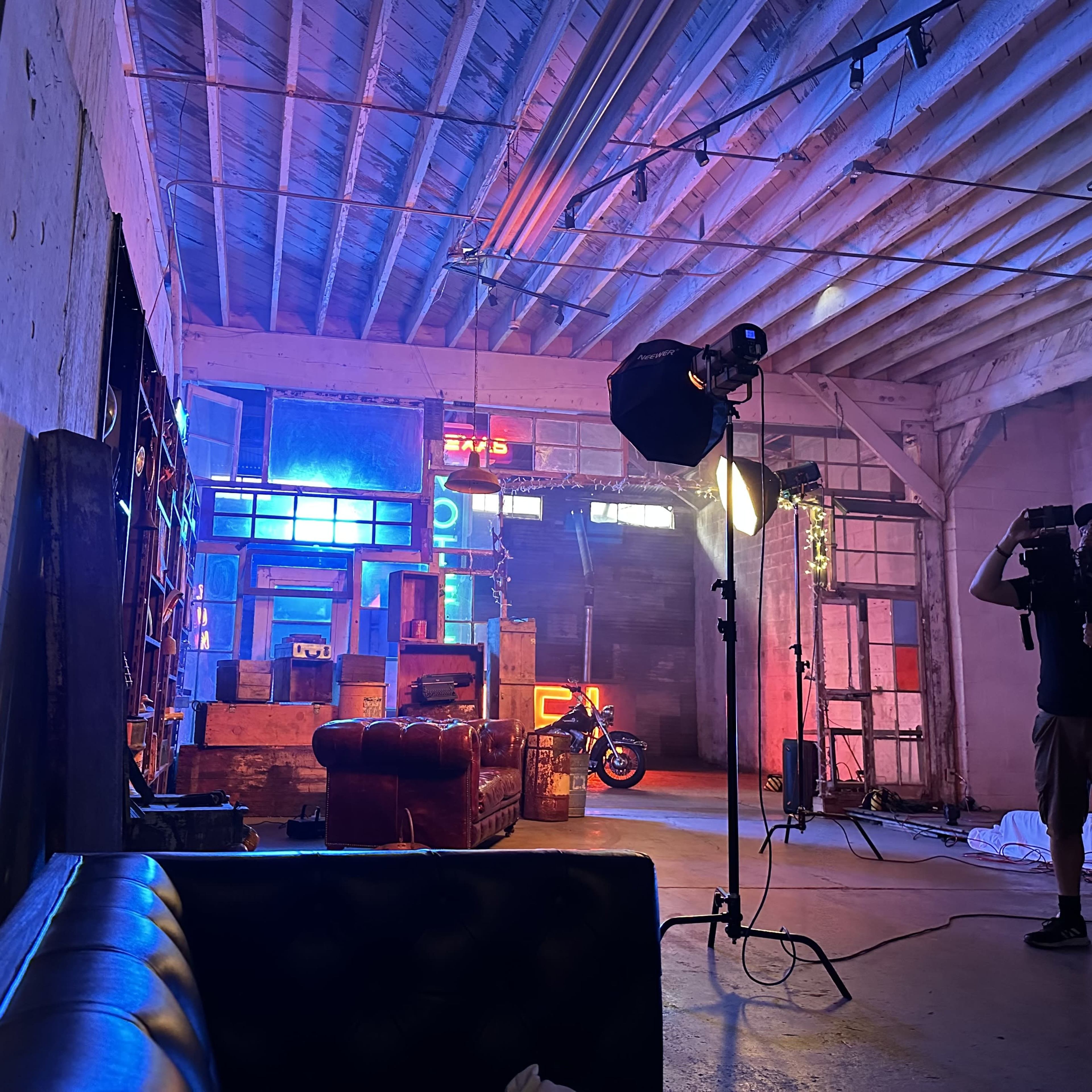 A photographer captures a brightly lit studio space featuring vintage furniture, neon signs, and a motorcycle in the background.
