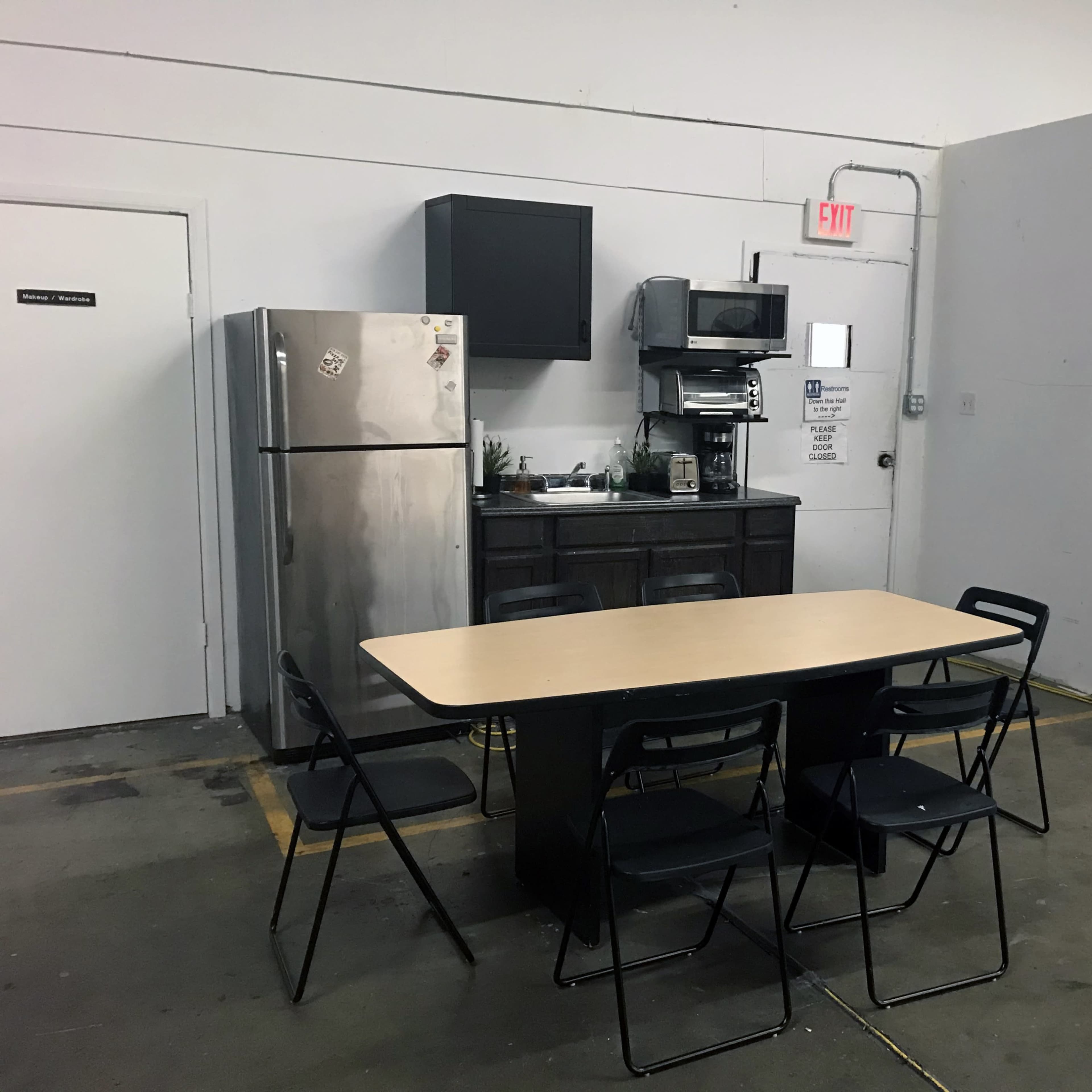 A simple dining area with a wooden table surrounded by six black chairs, adjacent to a small kitchen featuring a stainless steel refrigerator and a coffee maker.