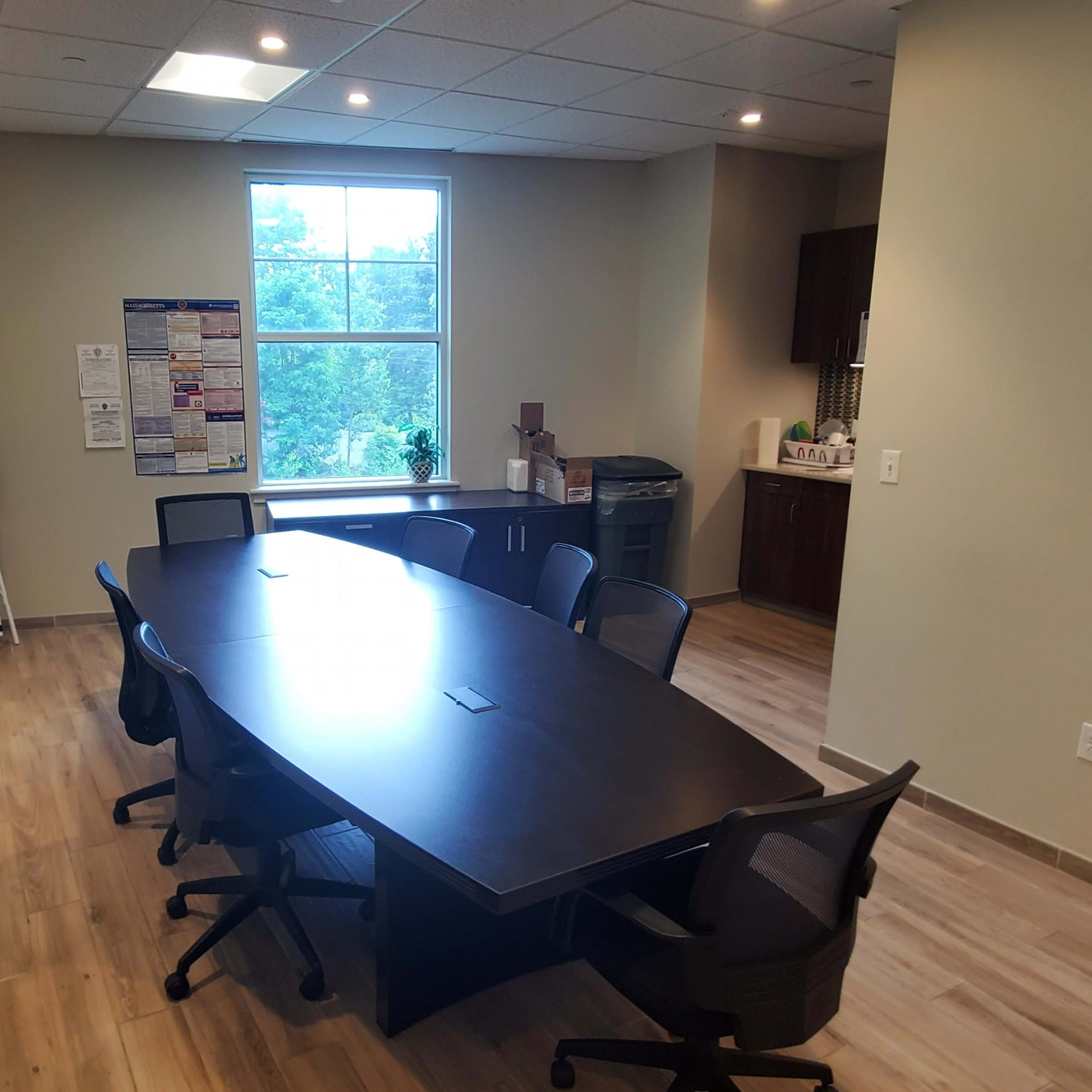 A conference room with a large table surrounded by several black chairs, adjacent to a kitchenette area and a restroom sign.