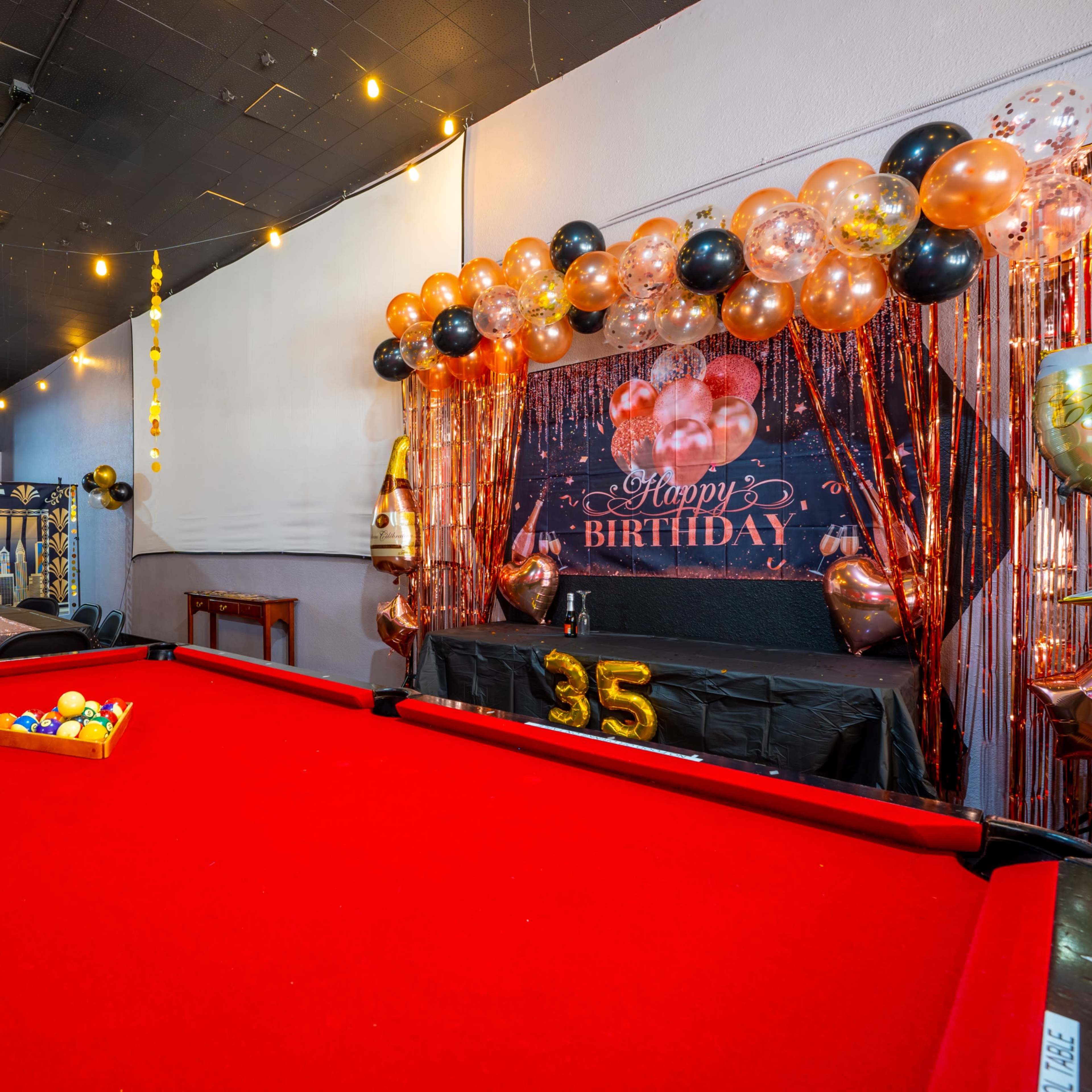 A red pool table is set up in a party room decorated with a black and gold birthday backdrop and elegant balloons for a 35th birthday celebration.
