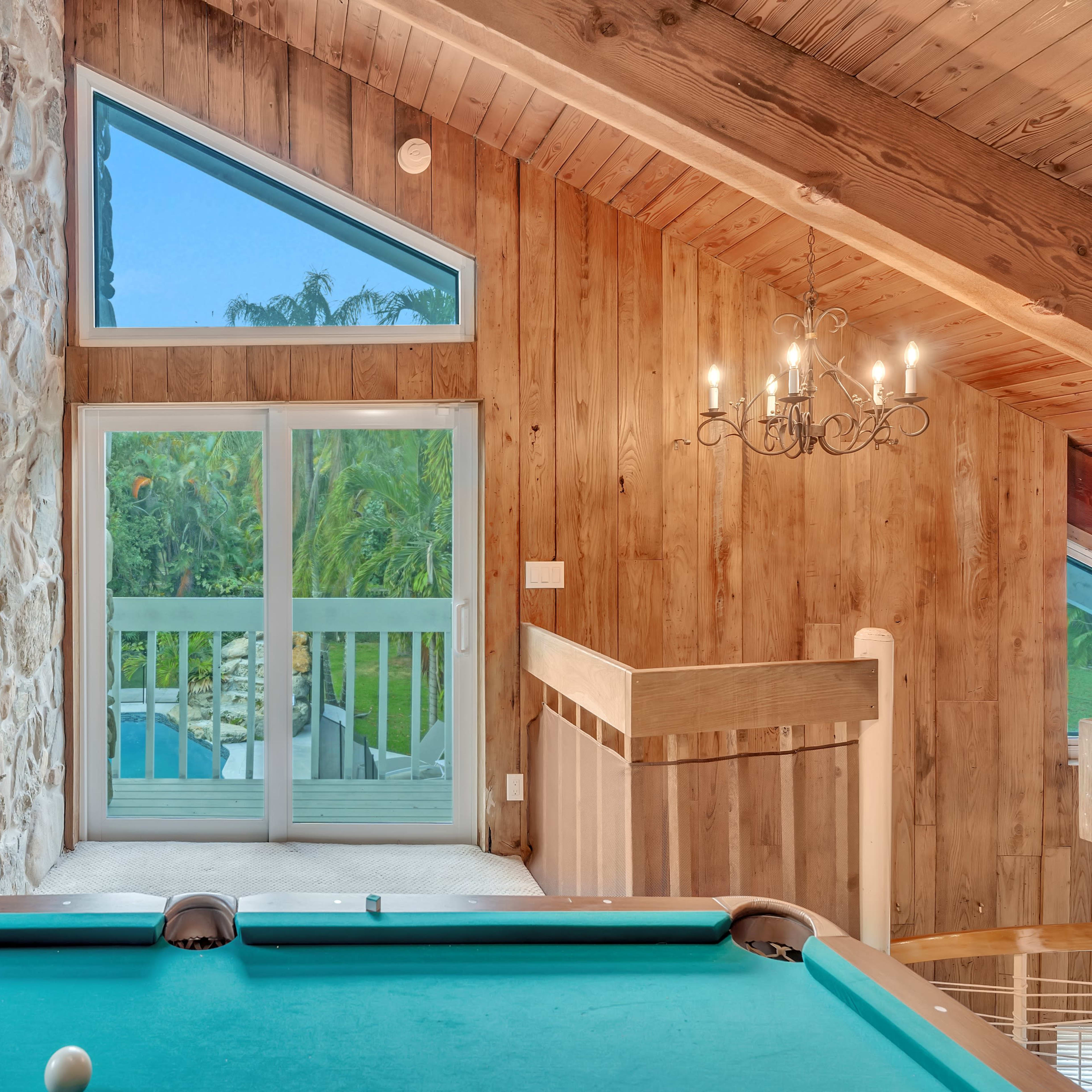 A spacious interior with a pool table in the foreground, large windows letting in natural light, and a view of greenery outside.
