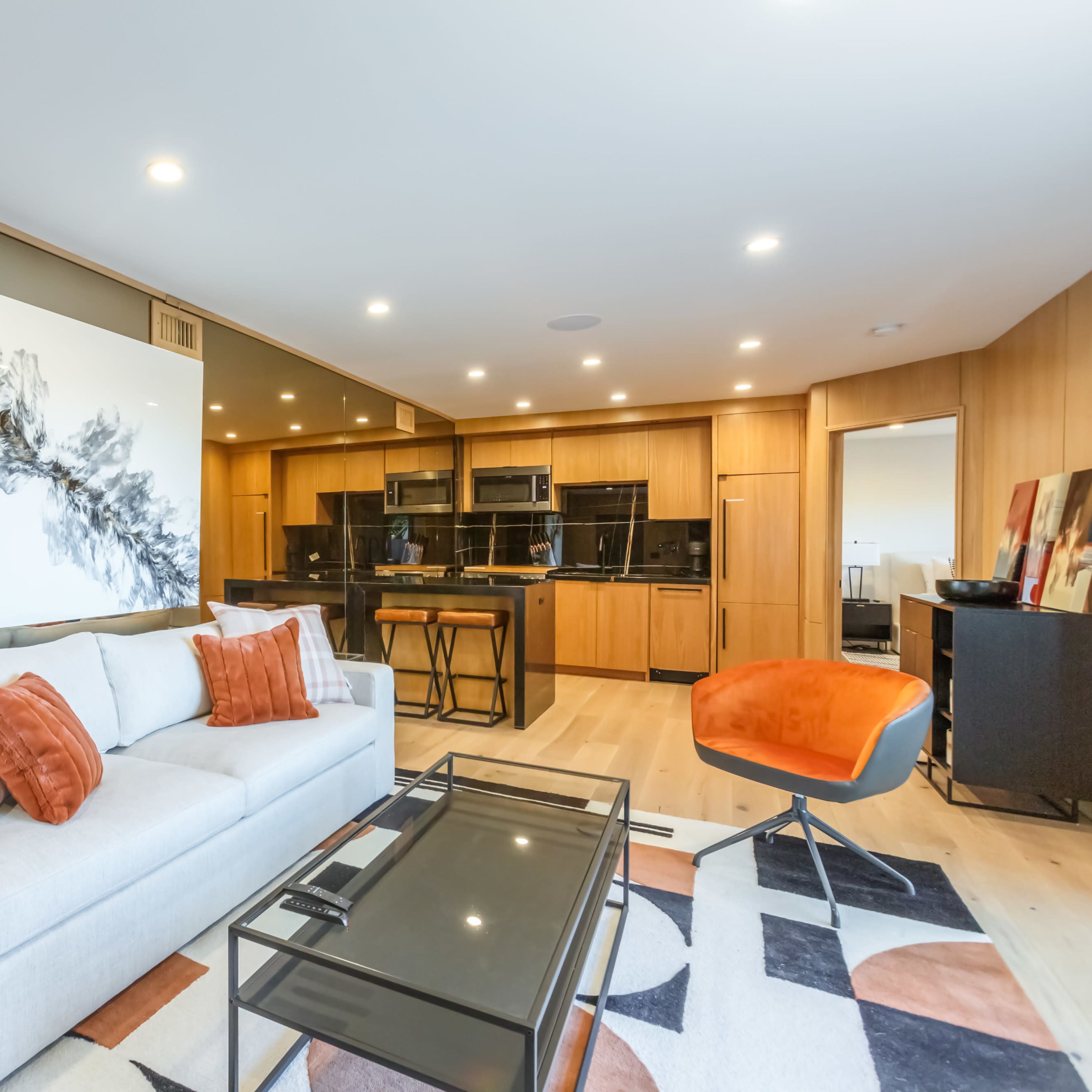 A modern living room featuring a light-colored sofa with orange and white pillows, a glass coffee table, and a stylish mural on the wall, with a dining area visible in the background.