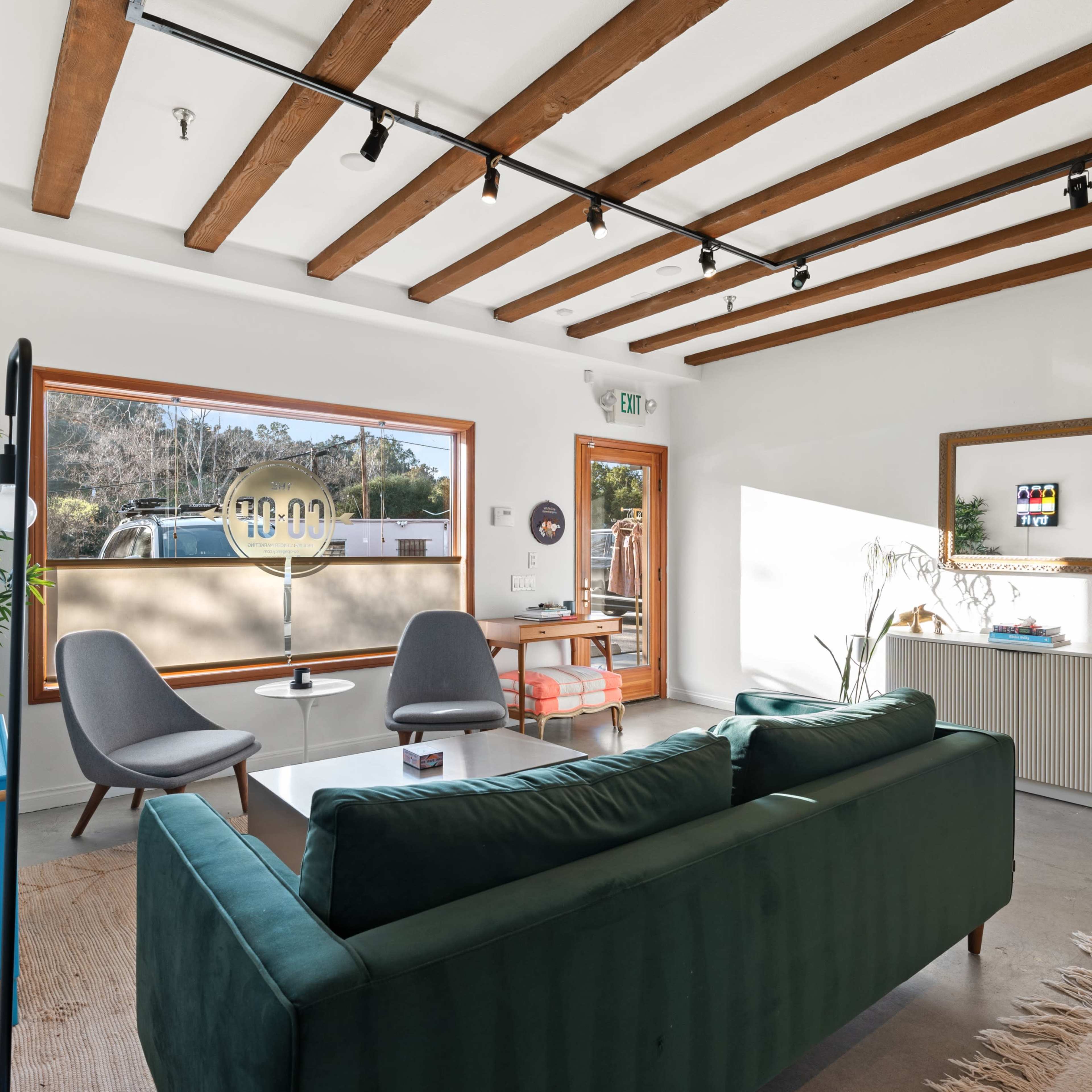 A modern living room features a green sofa, two gray chairs, a coffee table, and large windows that provide natural light.