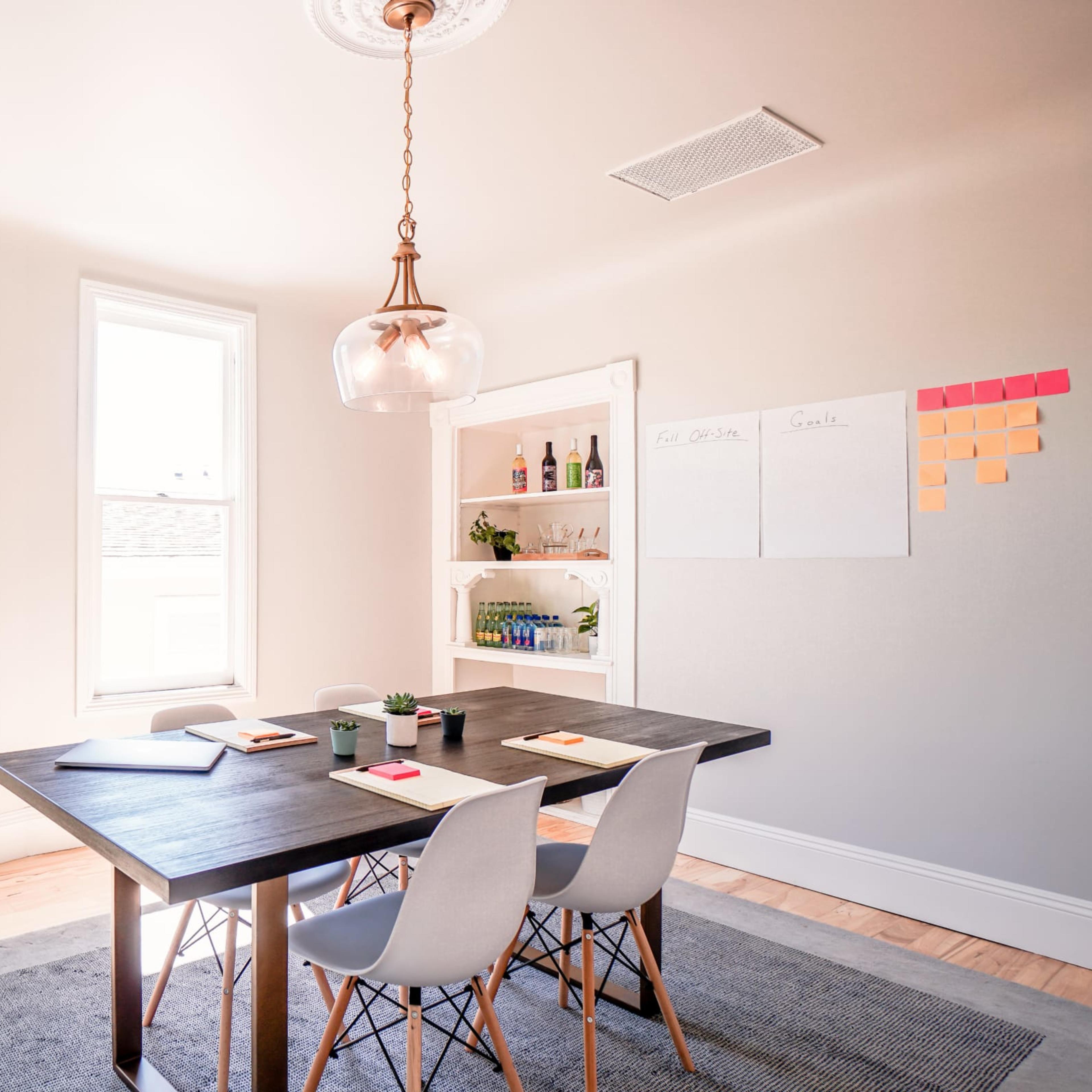 A bright, minimalist meeting room with a wooden table, four chairs, and a shelf containing bottles, alongside a wall featuring notes and sticky papers.