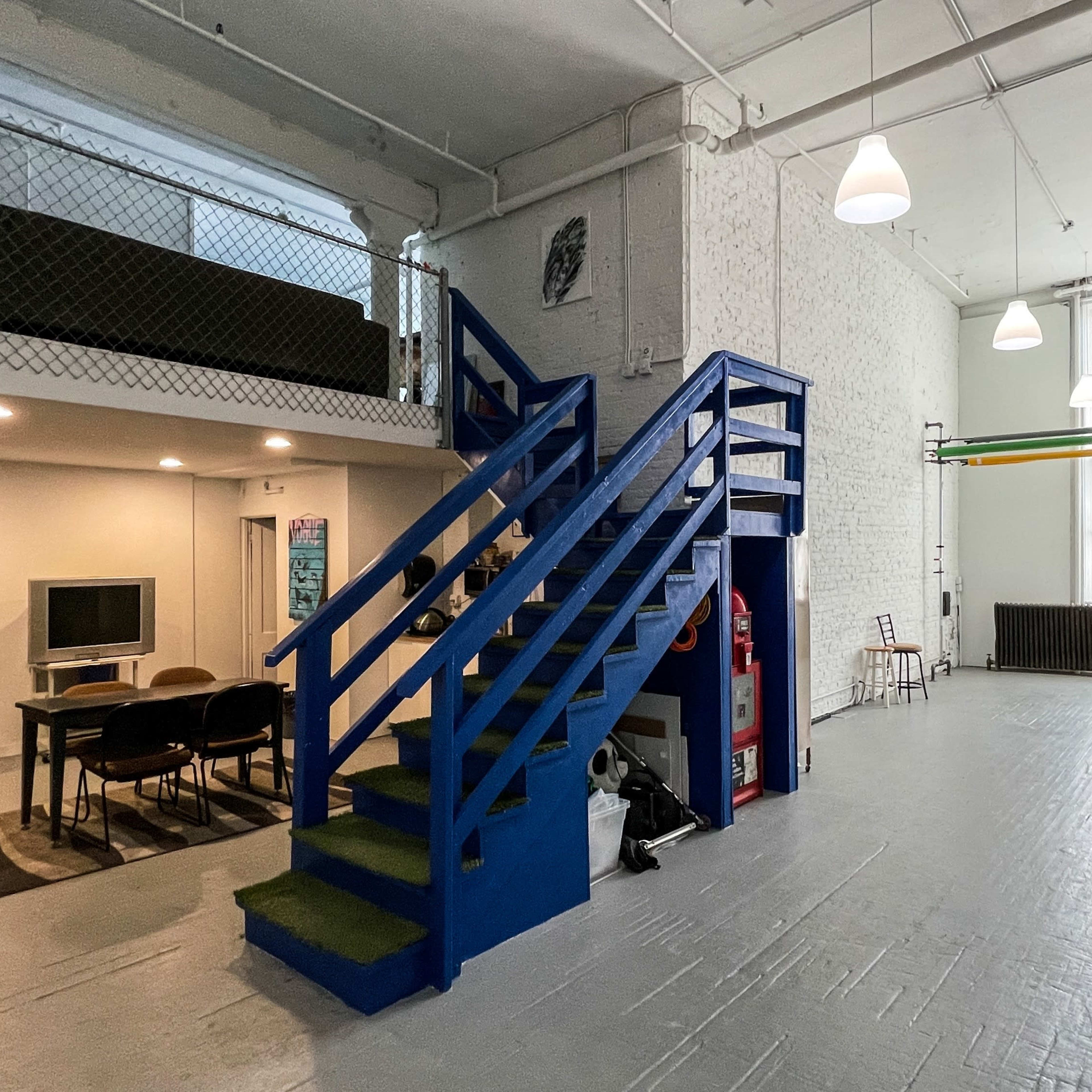 An open, industrial-style space featuring a staircase leading to a mezzanine, a seating area with a table and chairs, and large windows allowing natural light to enter.