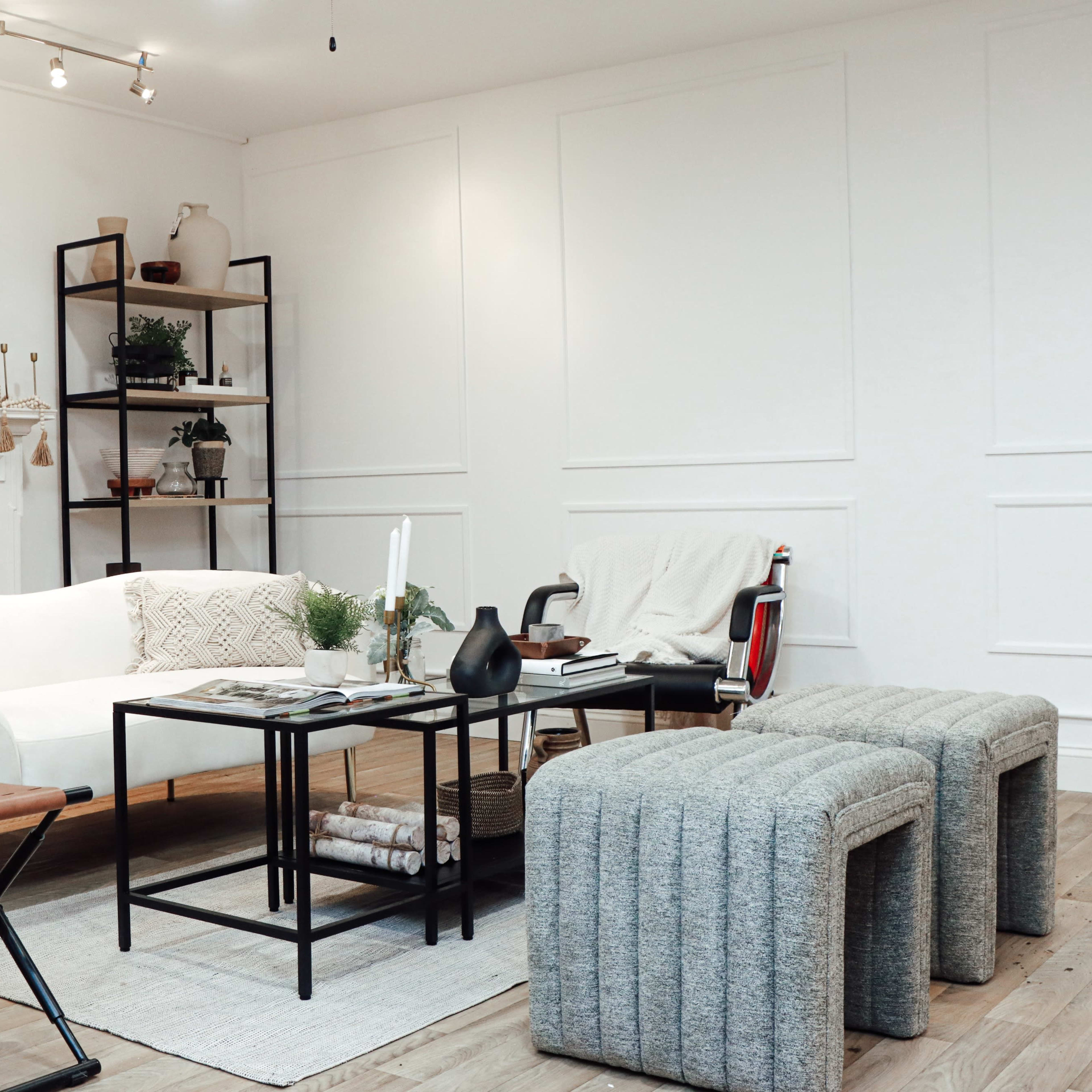 A modern living room with a white couch, a coffee table, two textured stools, and a bookshelf against a light-colored wall.