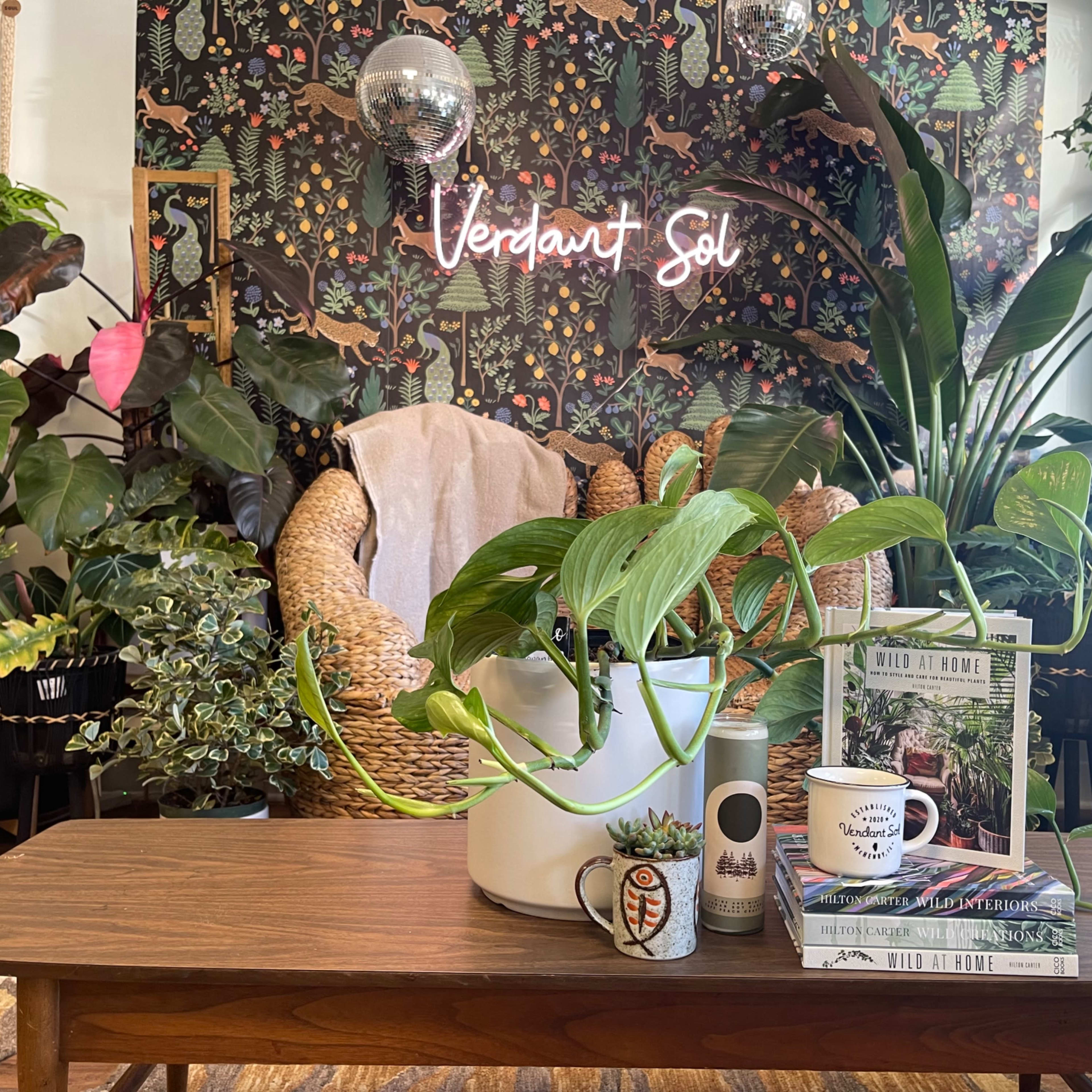 A cozy indoor space with a wooden table, a large potted plant, and a variety of greenery against a patterned wallpaper, highlighted by a neon sign that reads "Verdant Sol."