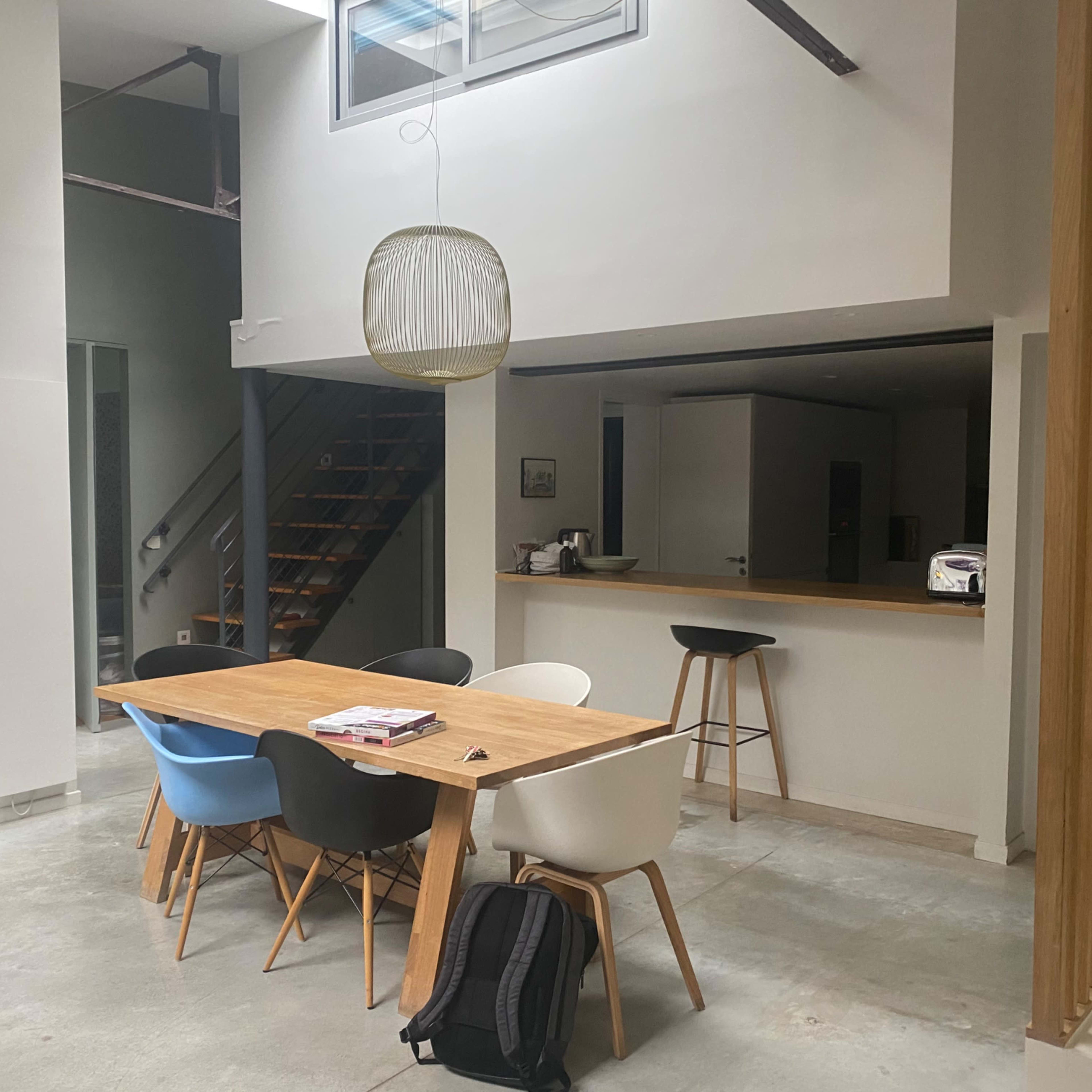 A modern, open-concept dining area with a wooden table surrounded by mismatched chairs, leading to a staircase and kitchen area.