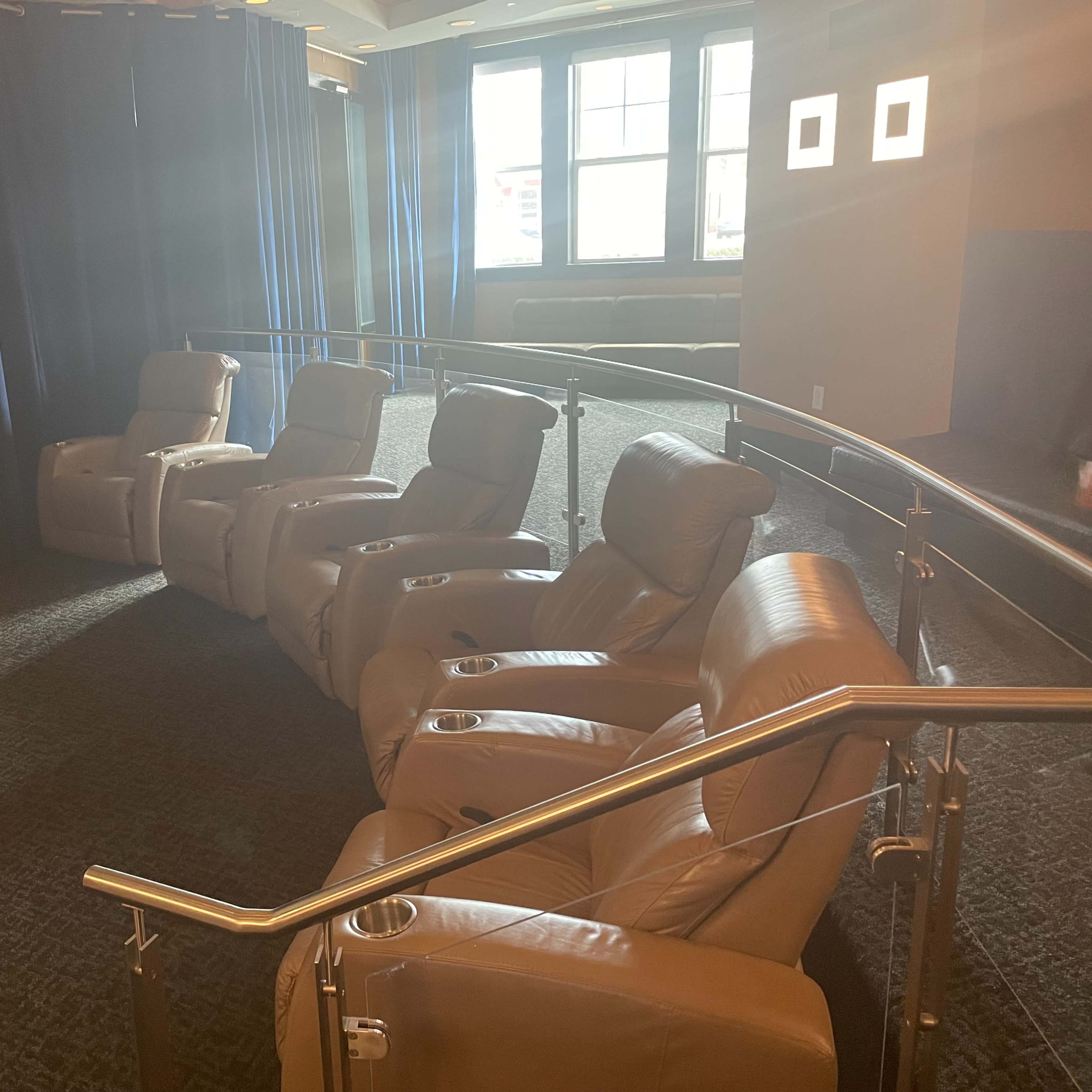 A row of beige reclining seats with cup holders, arranged on a carpeted floor in a room with blue curtains and windows in the background.