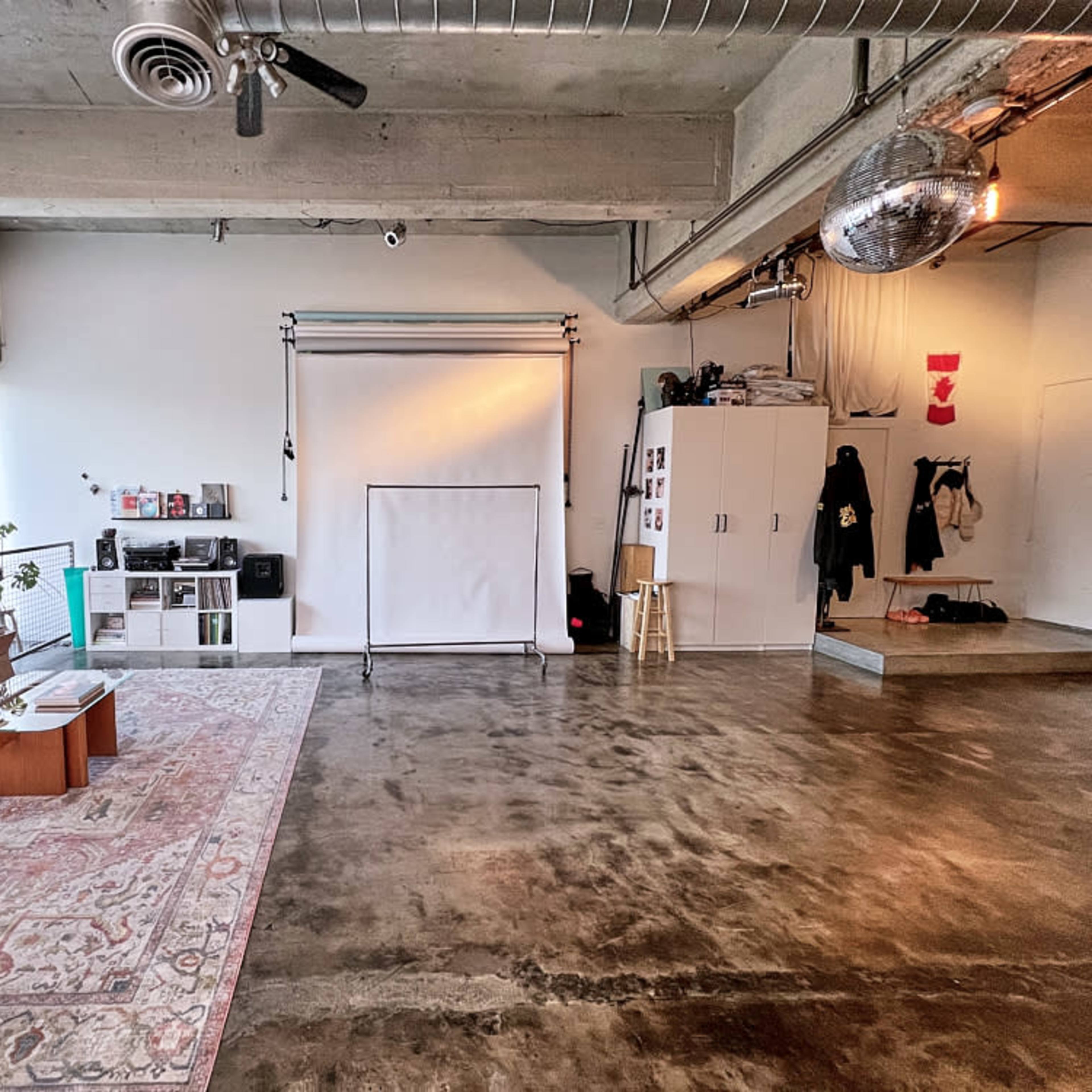 A spacious, industrial-style room with polished concrete floors, a seating area with a couch, a backdrop setup for photography, and storage cabinets along the wall.
