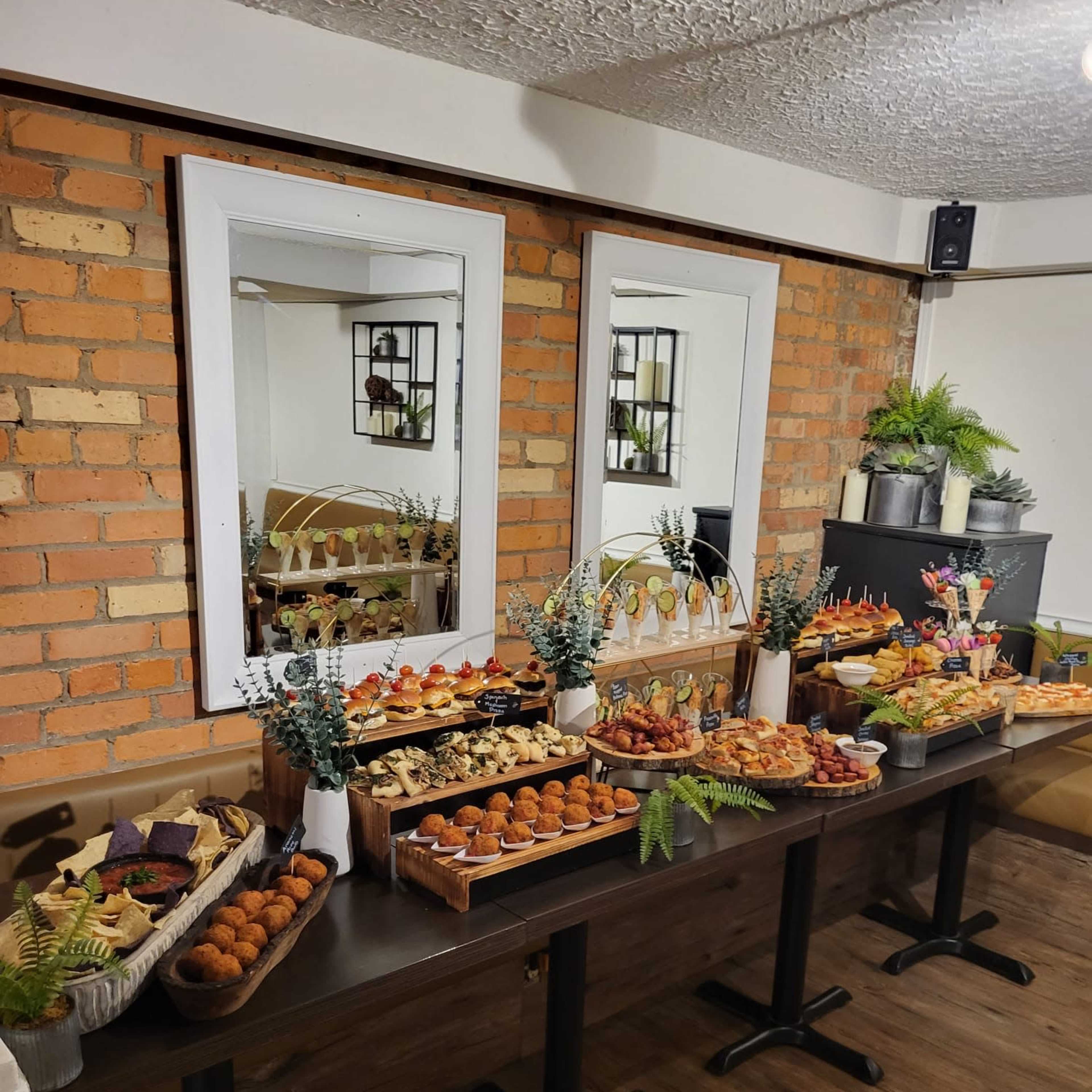 A long table is set with various dishes, including pastries, appetizers, and desserts, against a backdrop of brick walls and mirrors.
