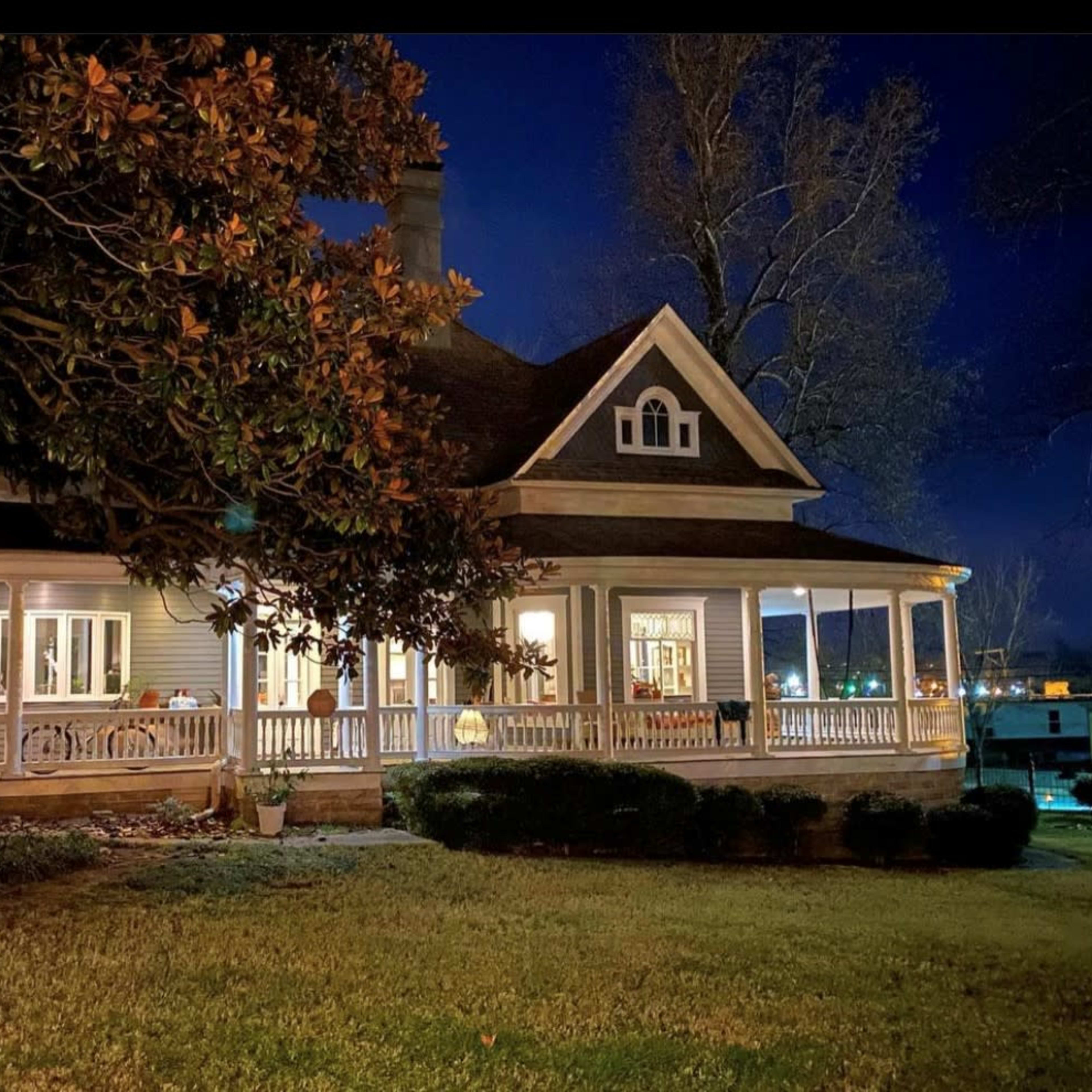 A two-story house with a wraparound porch and large windows is illuminated at night, surrounded by grass and trees.