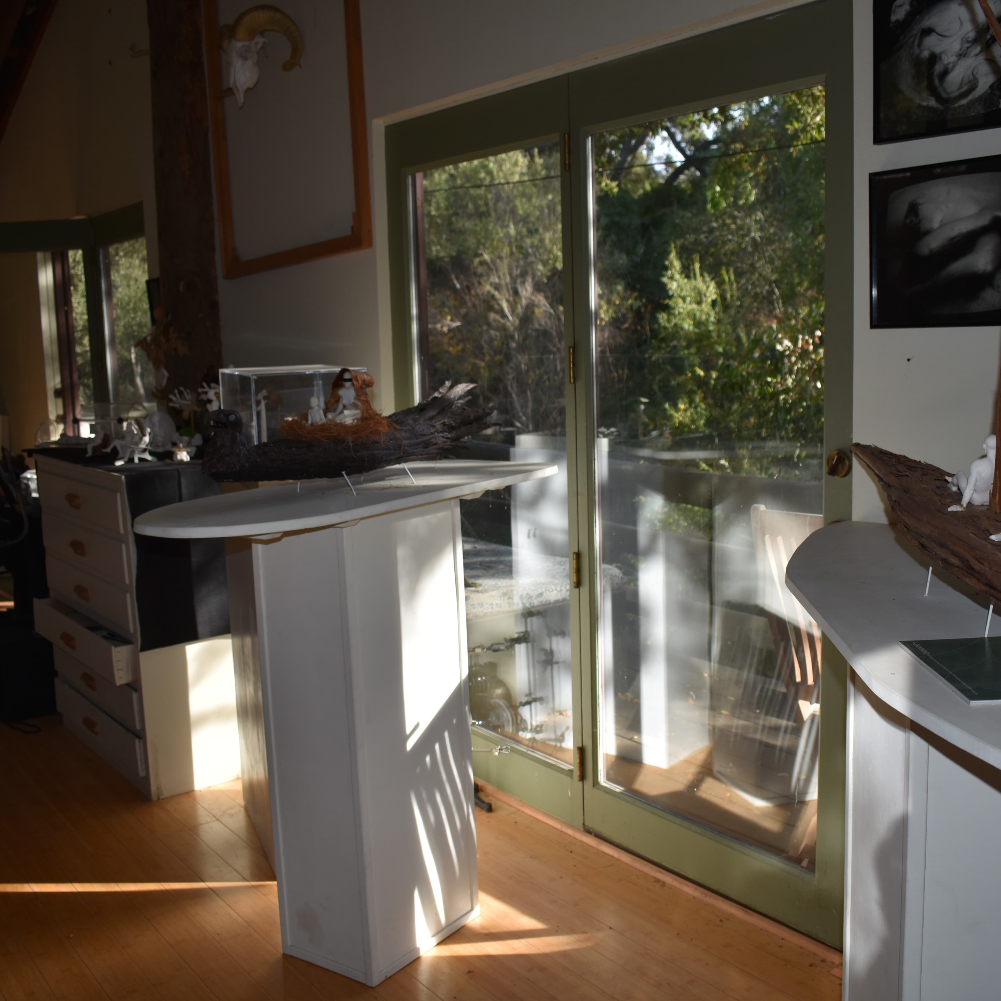 An indoor art studio with a wooden floor, featuring a table on the left and two sculptural pieces displayed on pedestals near a large window overlooking greenery.