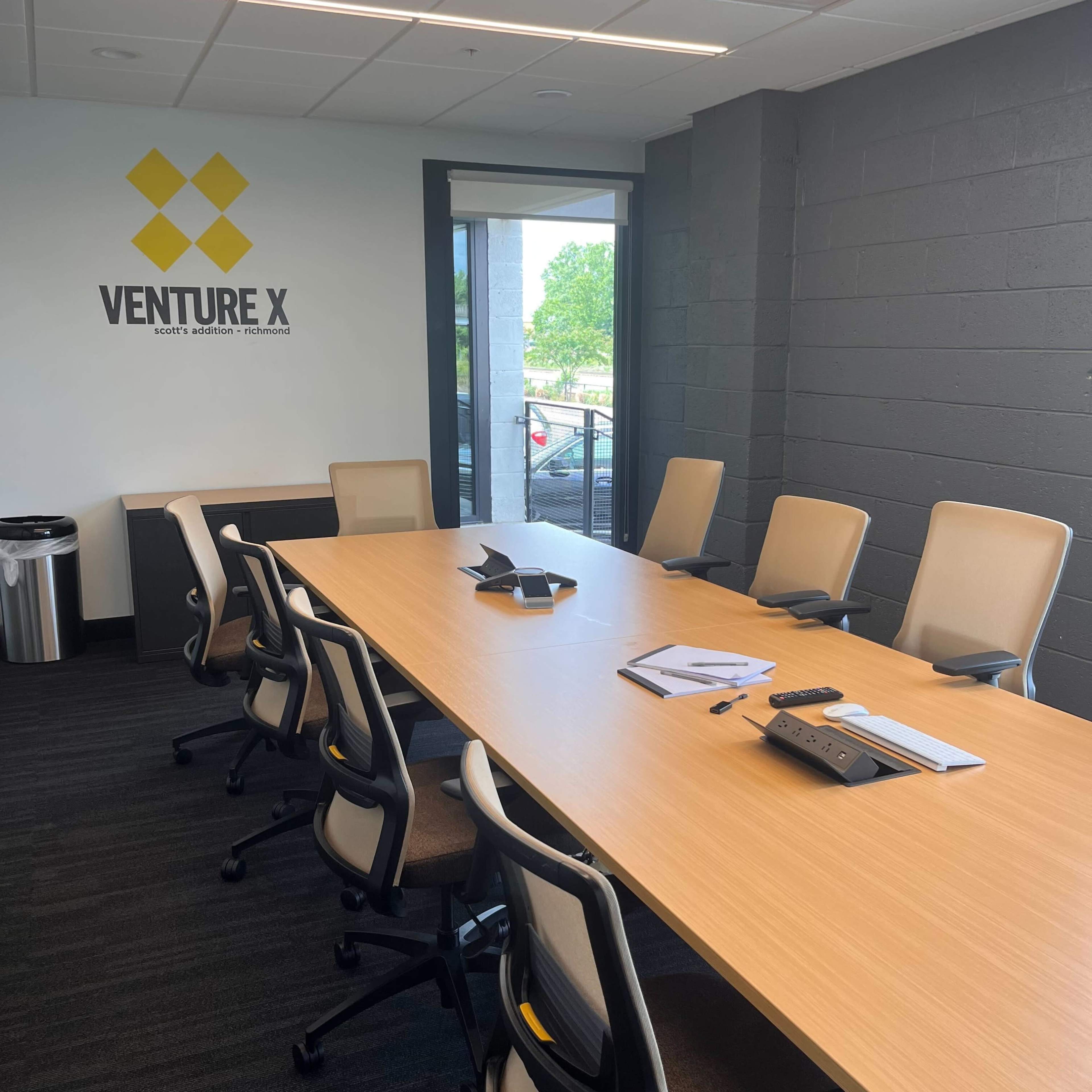 A spacious conference room features a long wooden table surrounded by ergonomic chairs, with a wall displaying the "Venture X" logo.