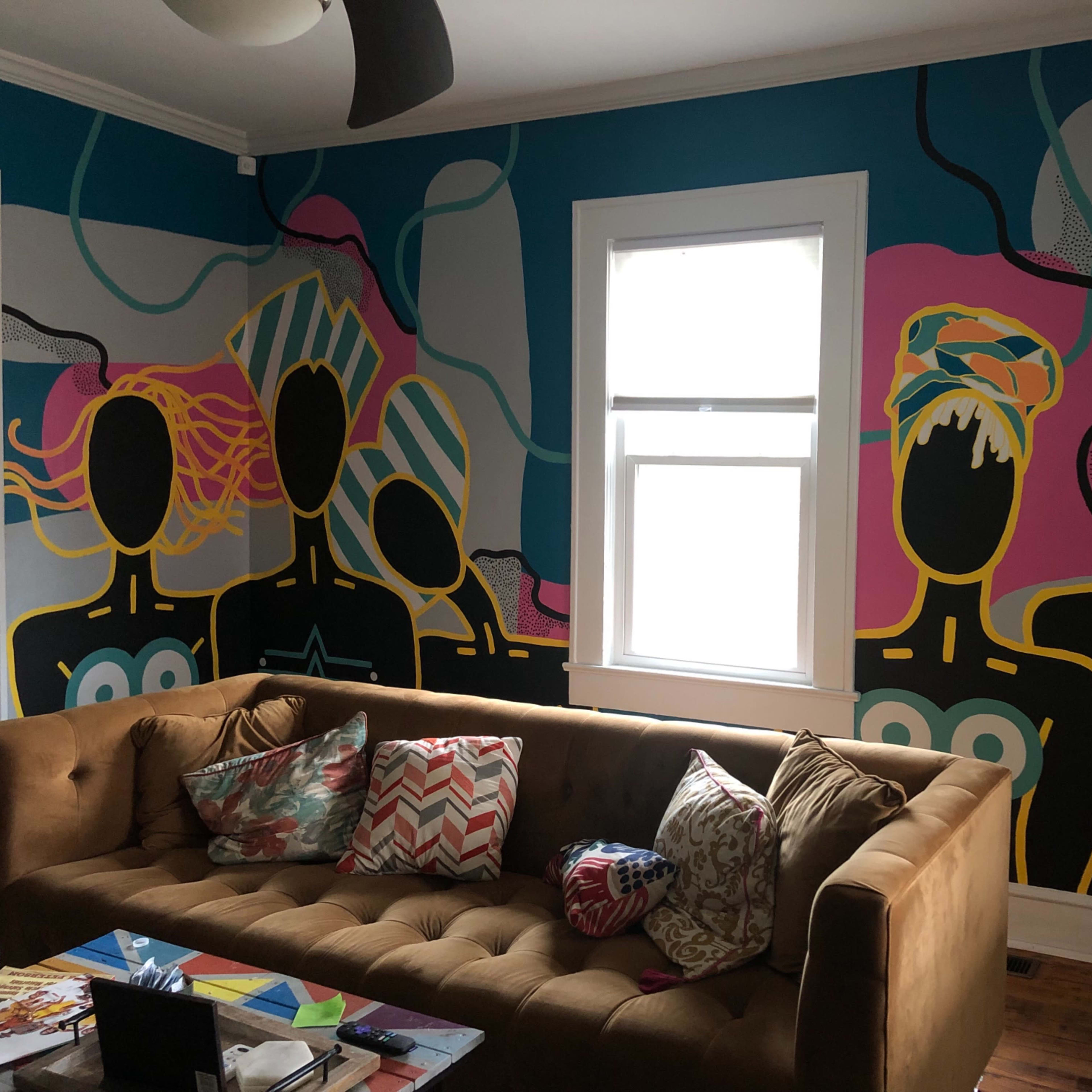 A living room with a brown sofa, colorful pillows, and a wall mural depicting three abstract figures with geometric patterns.