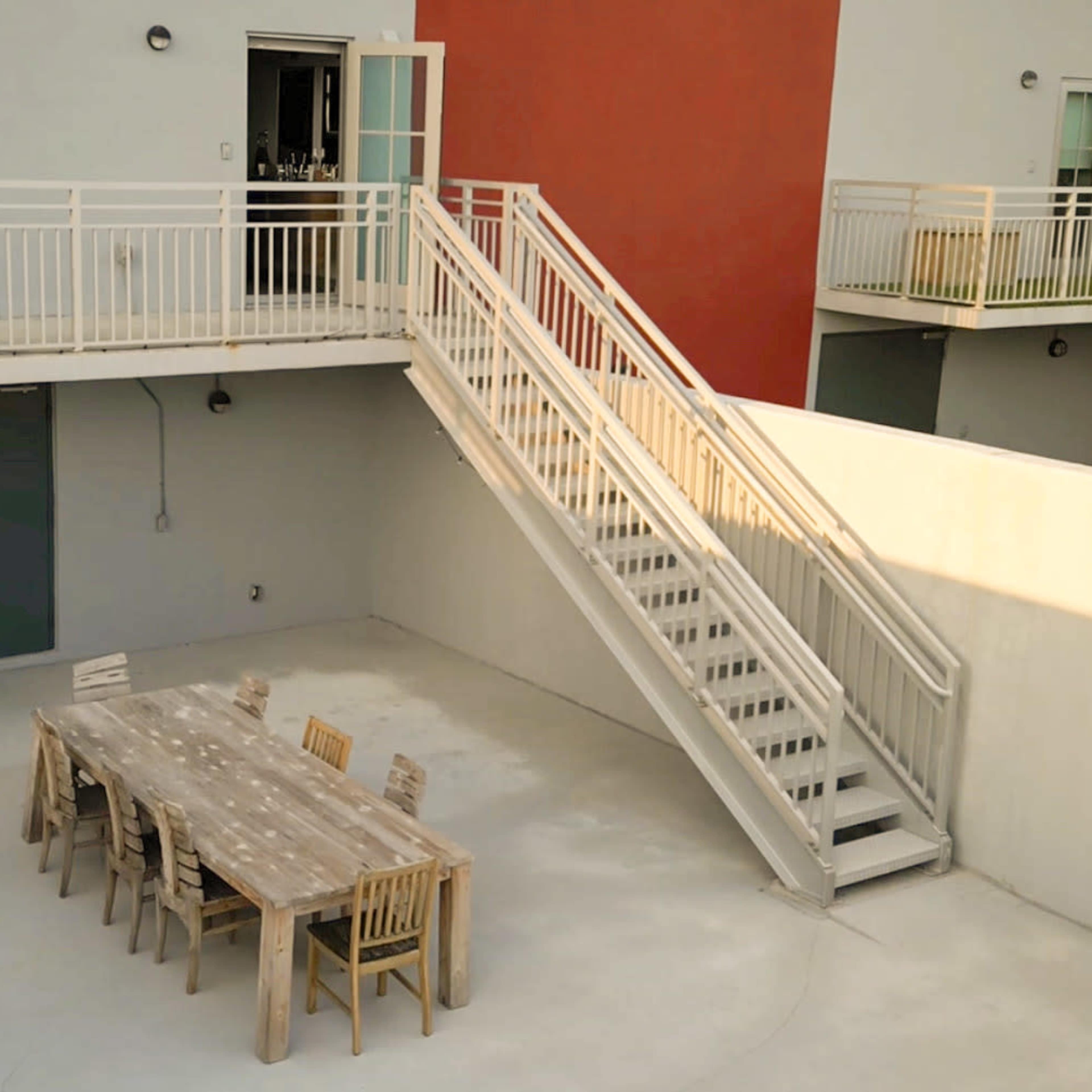 An outdoor courtyard featuring a long wooden table surrounded by chairs, with a flight of stairs leading to an upper level of a building.